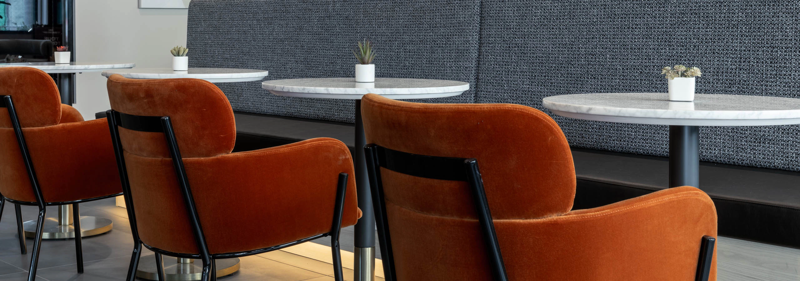 Social and co-working space with three round white tables, orange chairs, and long built-in bench seating opposite.