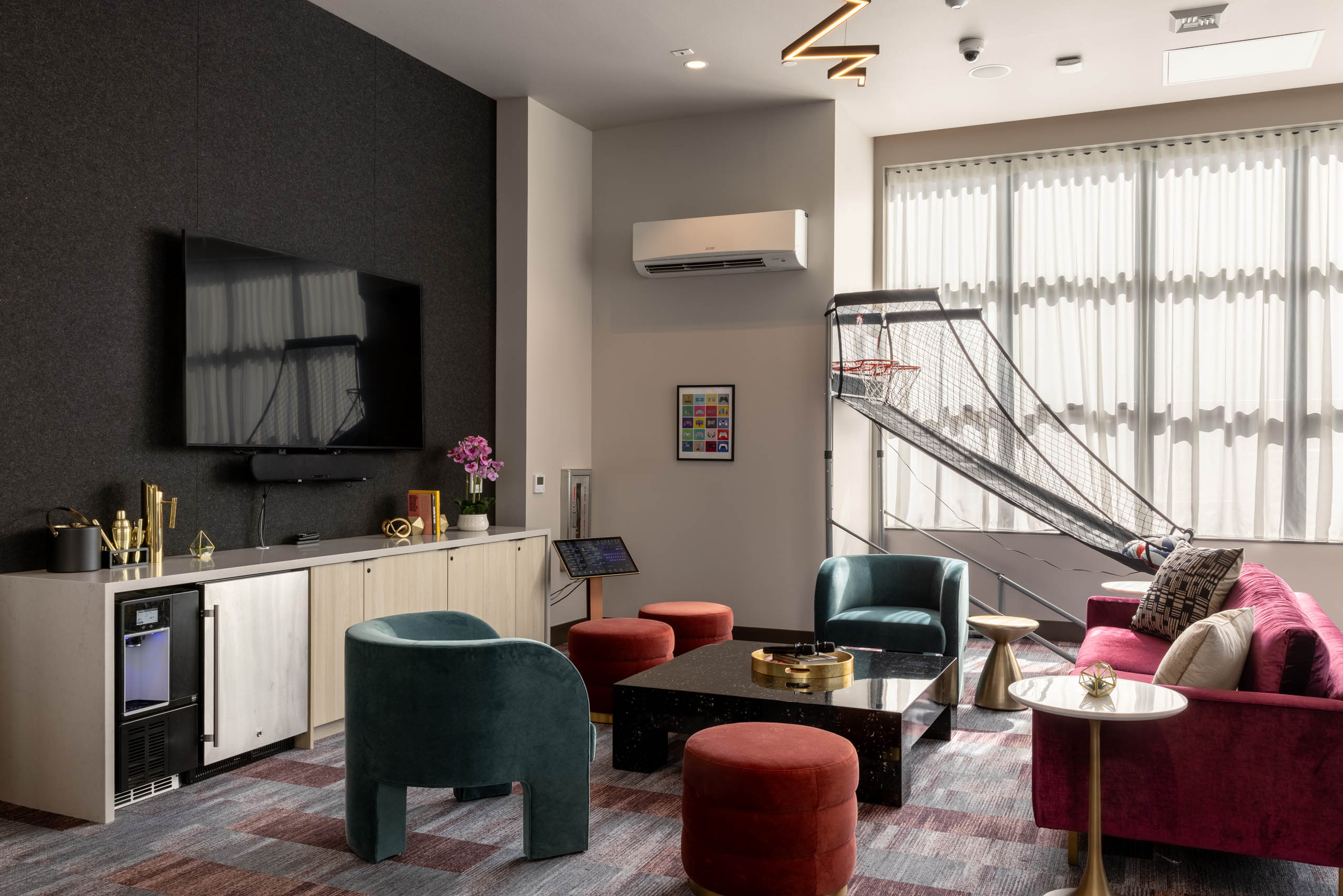 Clubroom with colorful seating, a wall-mounted TV, coffee table, and a basketball arcade game near the windows.