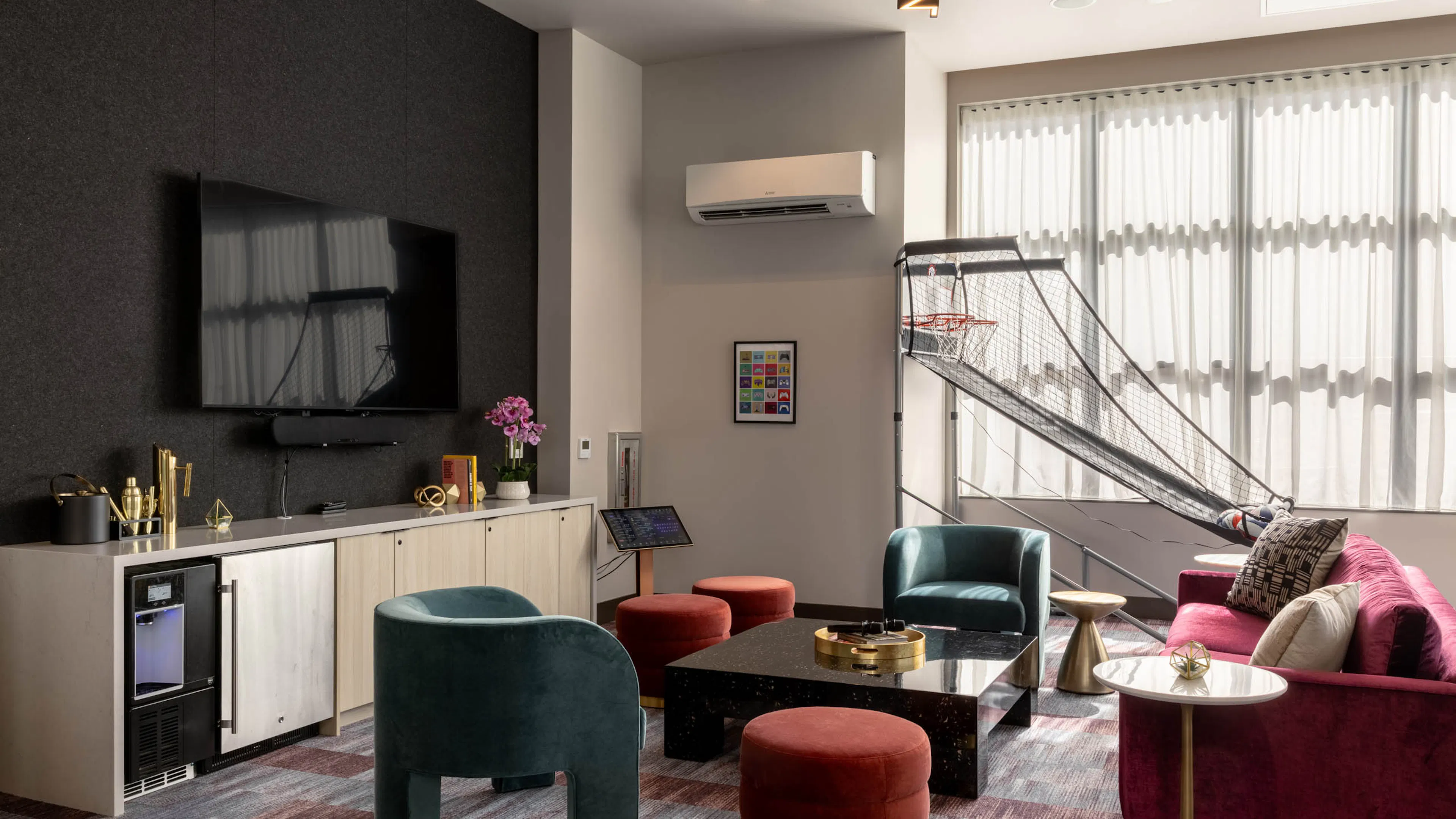 Clubroom with colorful seating, a wall-mounted TV, coffee table, and a basketball arcade game near the windows.