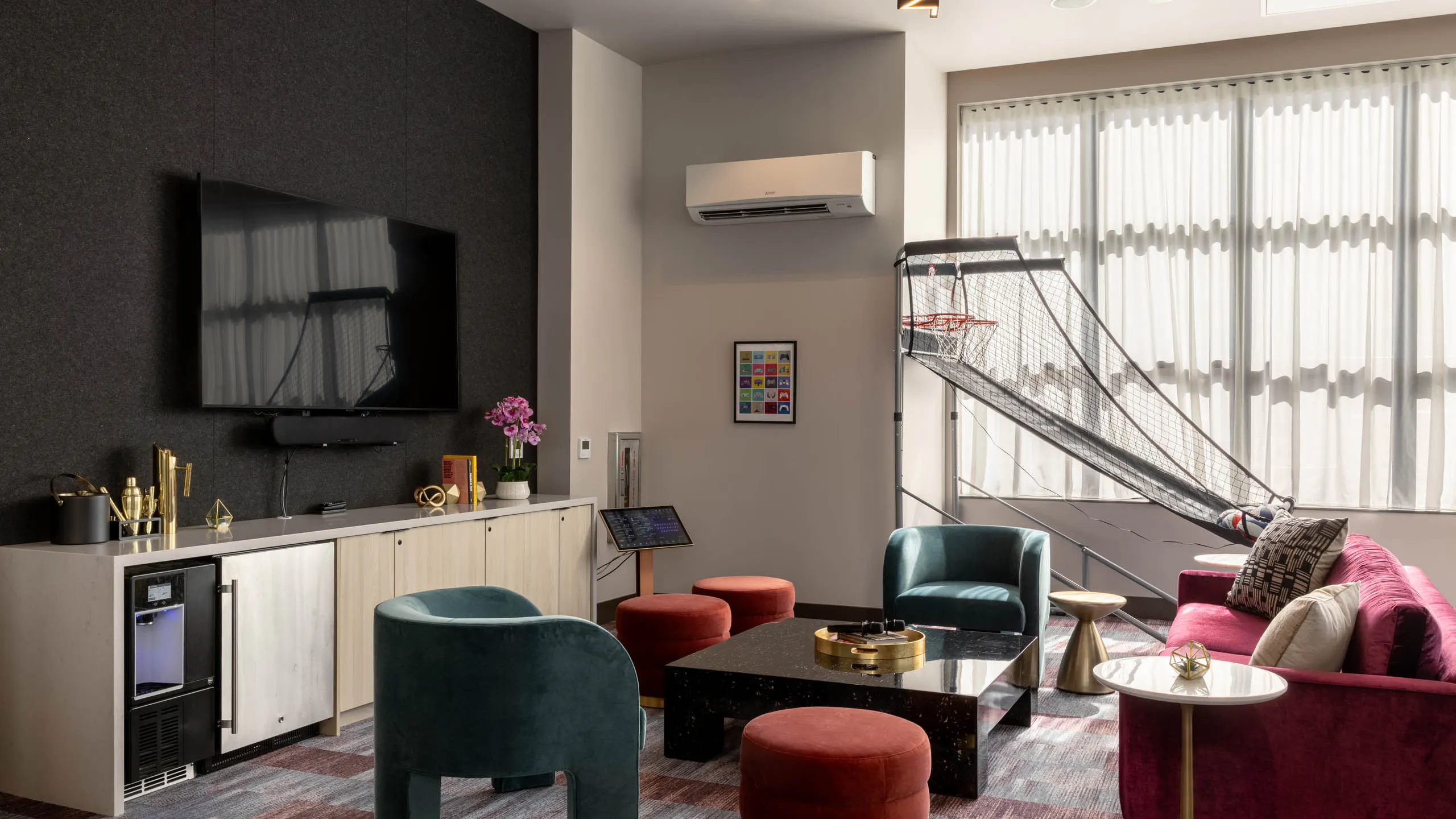 Clubroom with colorful seating, a wall-mounted TV, coffee table, and a basketball arcade game near the windows.