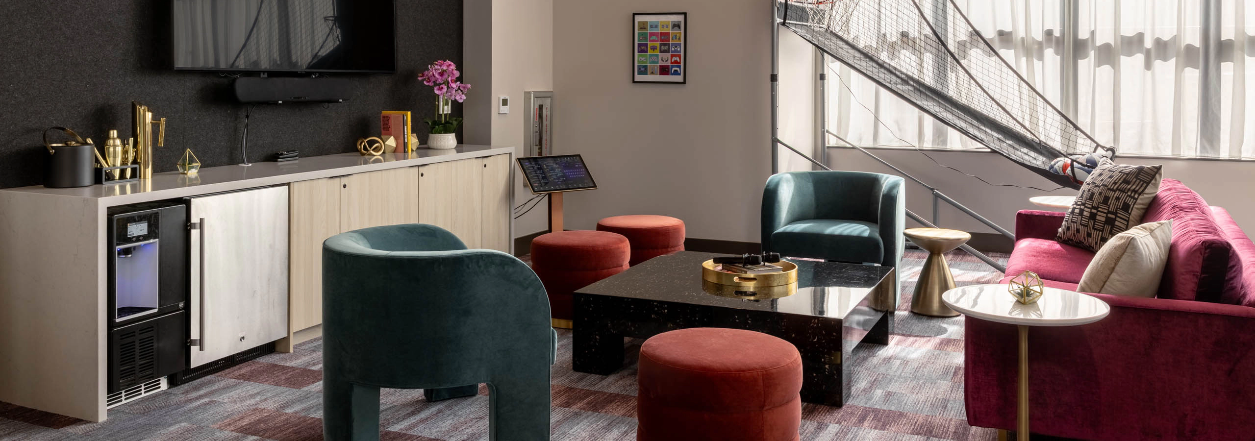 Clubroom with colorful seating, a wall-mounted TV, coffee table, and a basketball arcade game near the windows.
