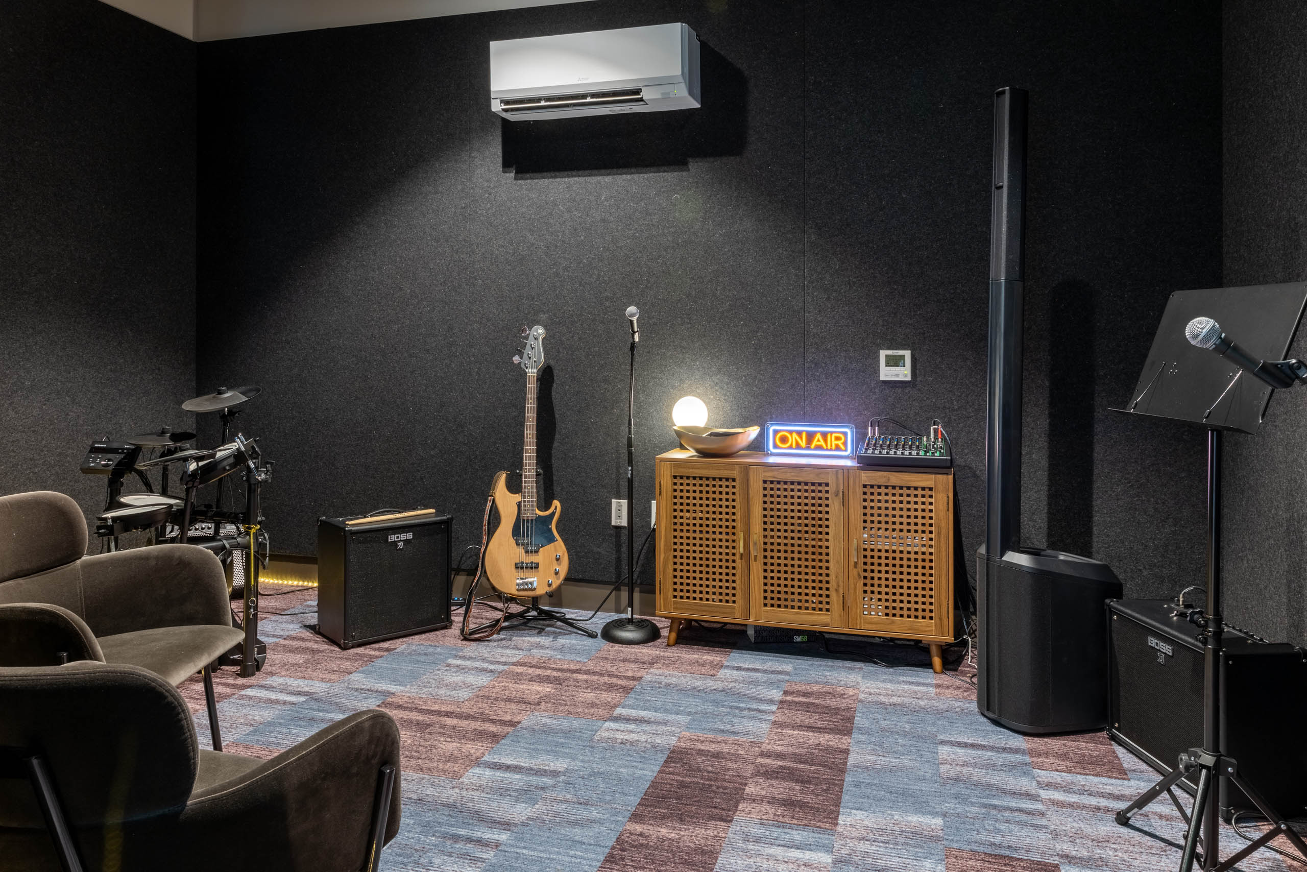 Music station in a soundproof room with two brown chairs, a microphone and stand, guitar, drum set, and amplifiers.