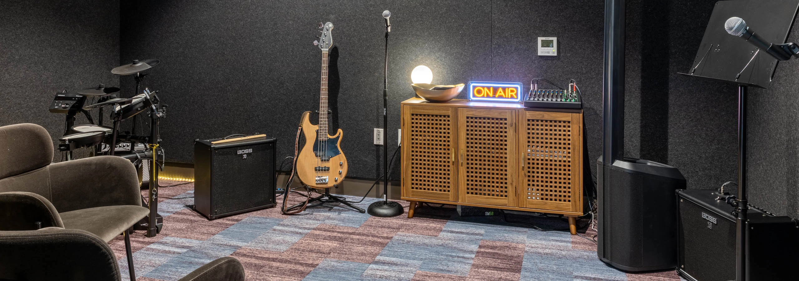 Music station in a soundproof room with two brown chairs, a microphone and stand, guitar, drum set, and amplifiers.