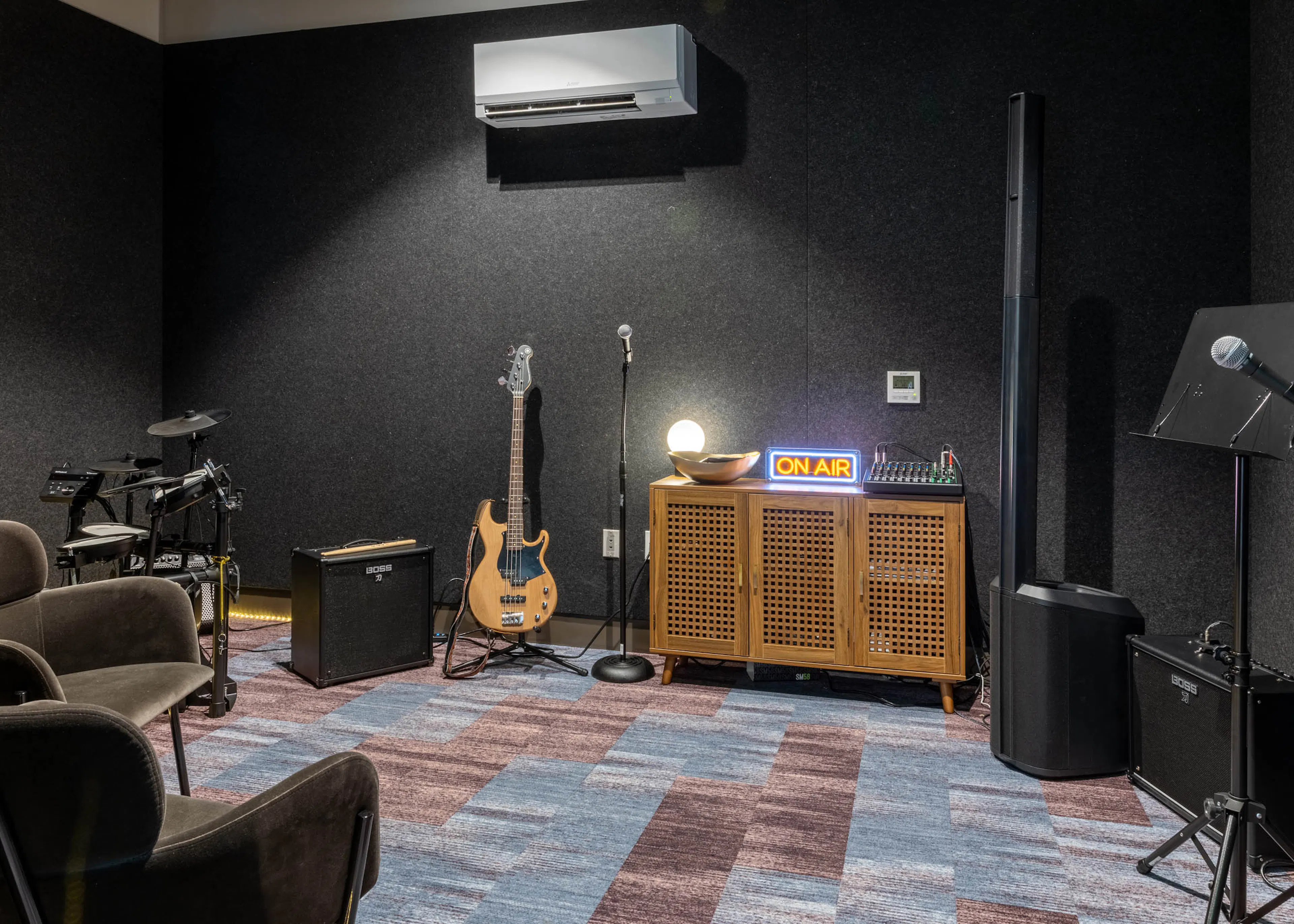 Music station in a soundproof room with two brown chairs, a microphone and stand, guitar, drum set, and amplifiers.