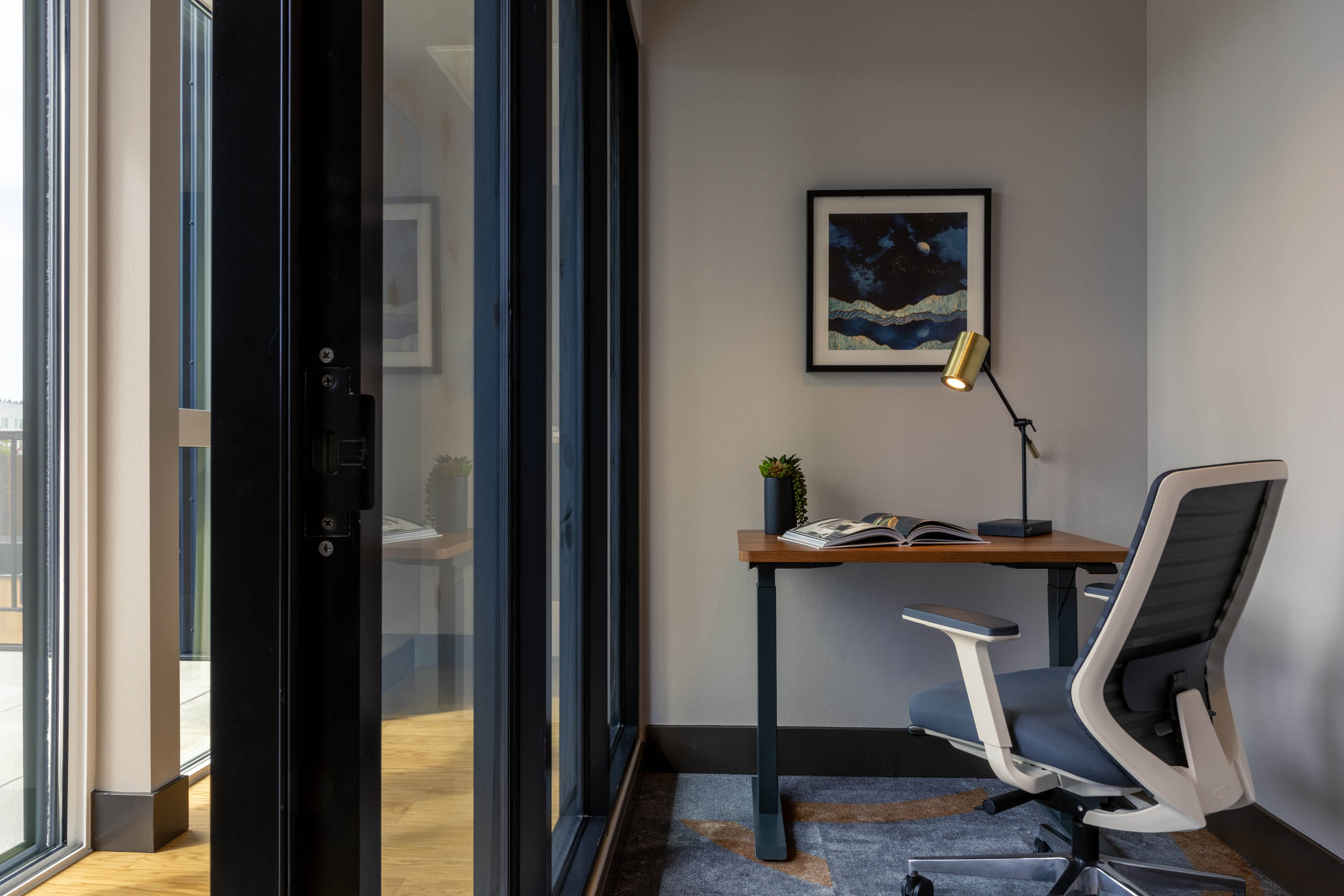 Work-from-home space with a wooden desk and chair, floor-to-ceiling windows, white walls, and blue carpet.