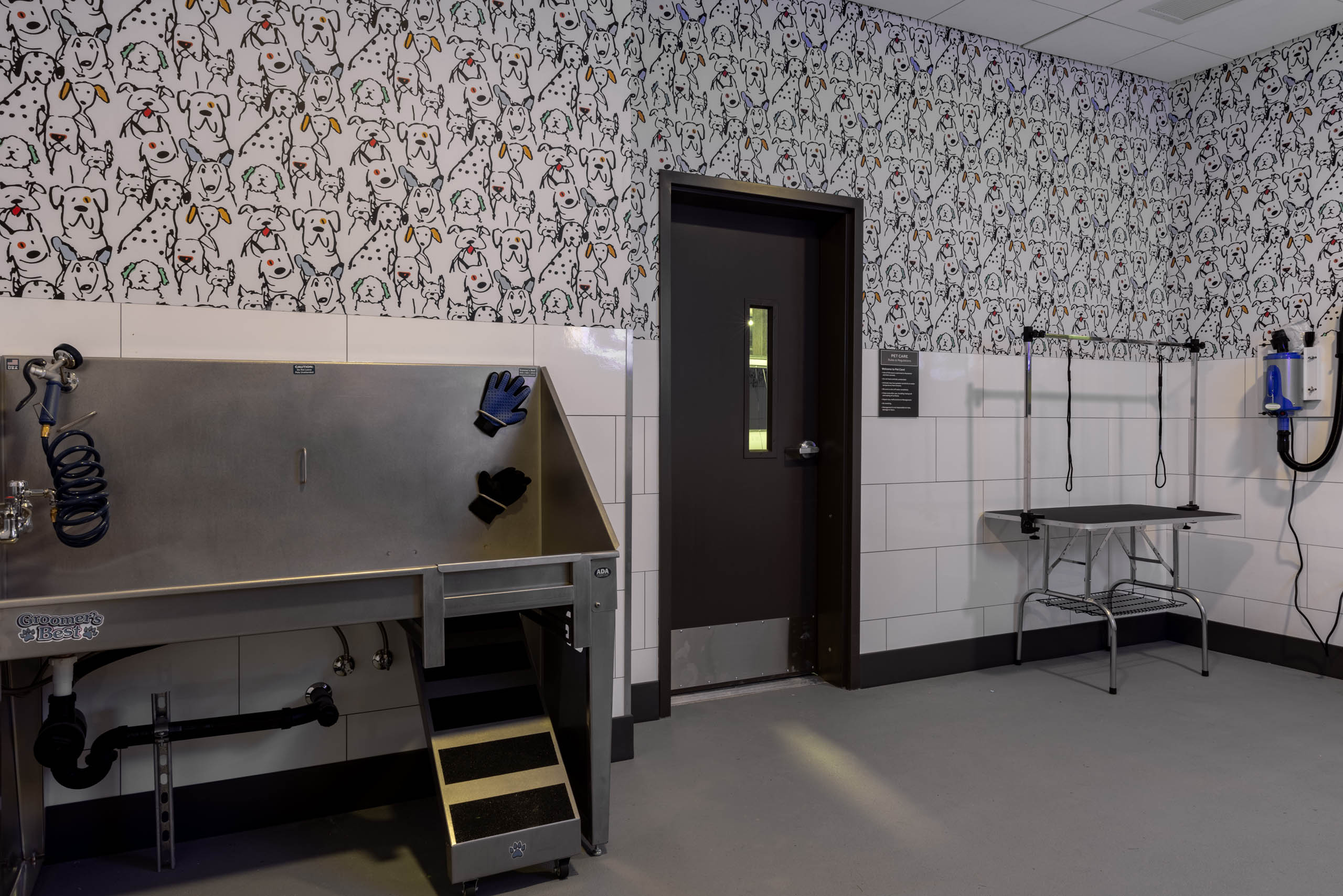 Pet wash station with stainless steel grooming tub, drying tools, and grooming table in a tiled room with dog-patterned wallpaper.