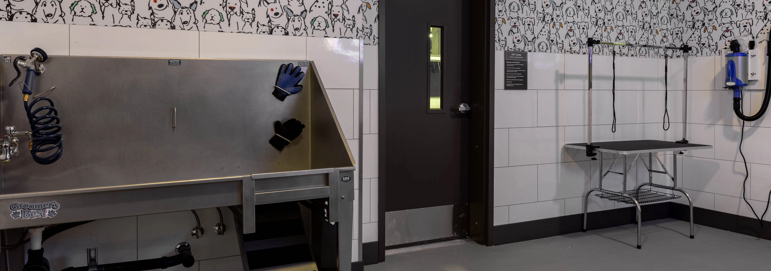 Pet wash station with stainless steel grooming tub, drying tools, and grooming table in a tiled room with dog-patterned wallpaper.