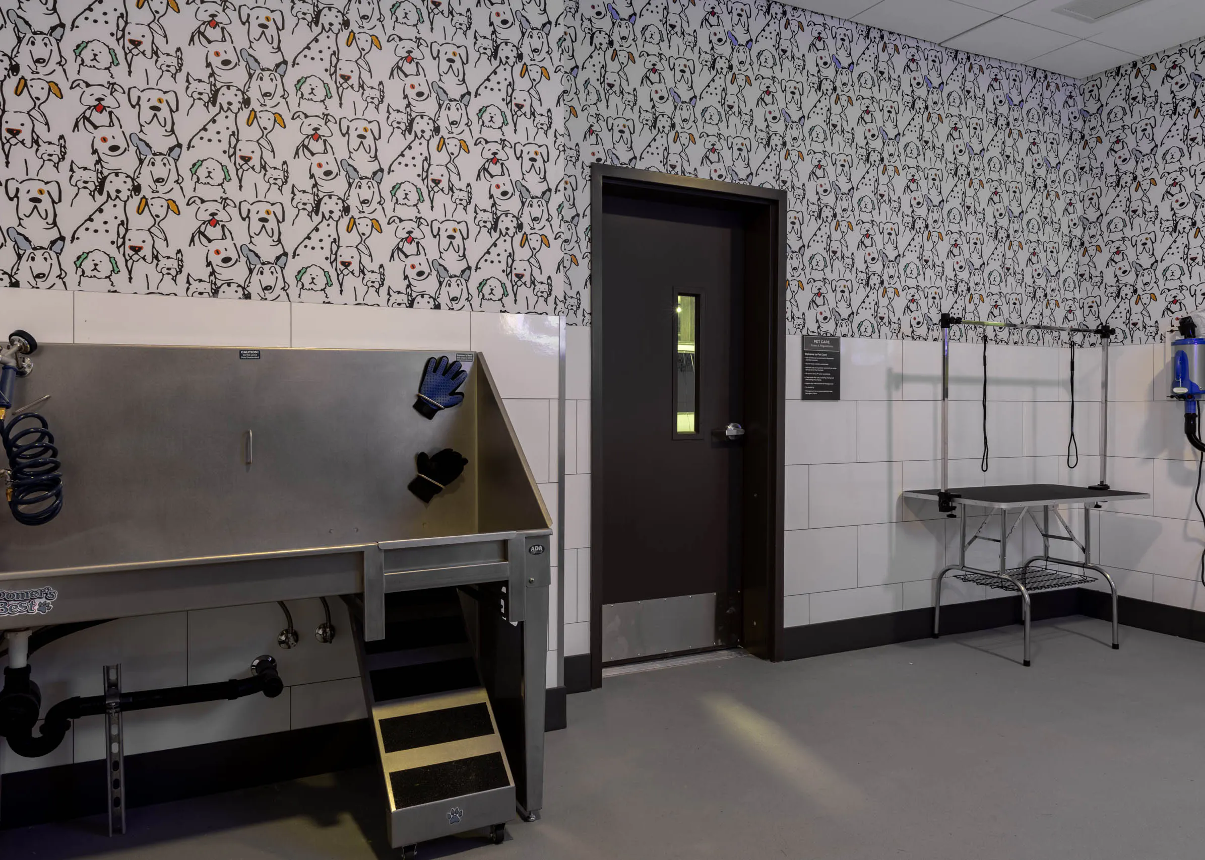 Pet wash station with stainless steel grooming tub, drying tools, and grooming table in a tiled room with dog-patterned wallpaper.