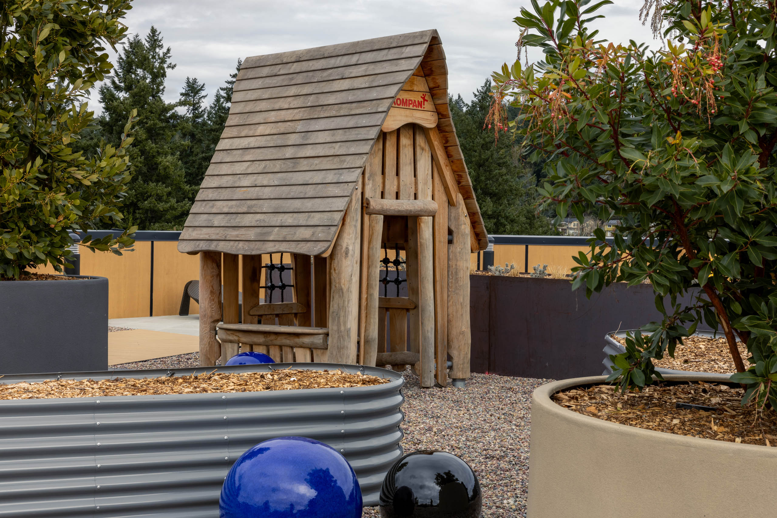 Play area for children with a wooden playhouse surrounded by two potted plants on a rooftop terrace.