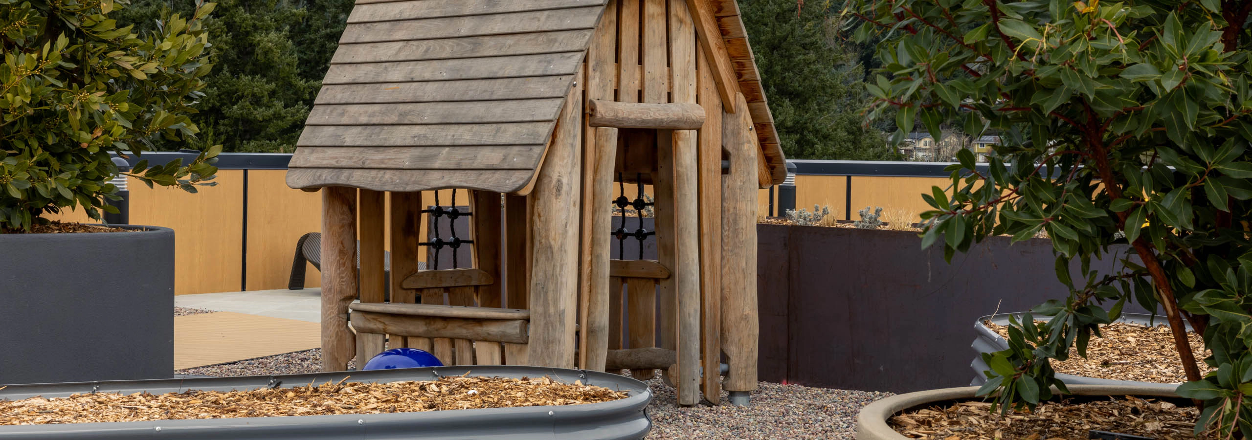 Play area for children with a wooden playhouse surrounded by two potted plants on a rooftop terrace.