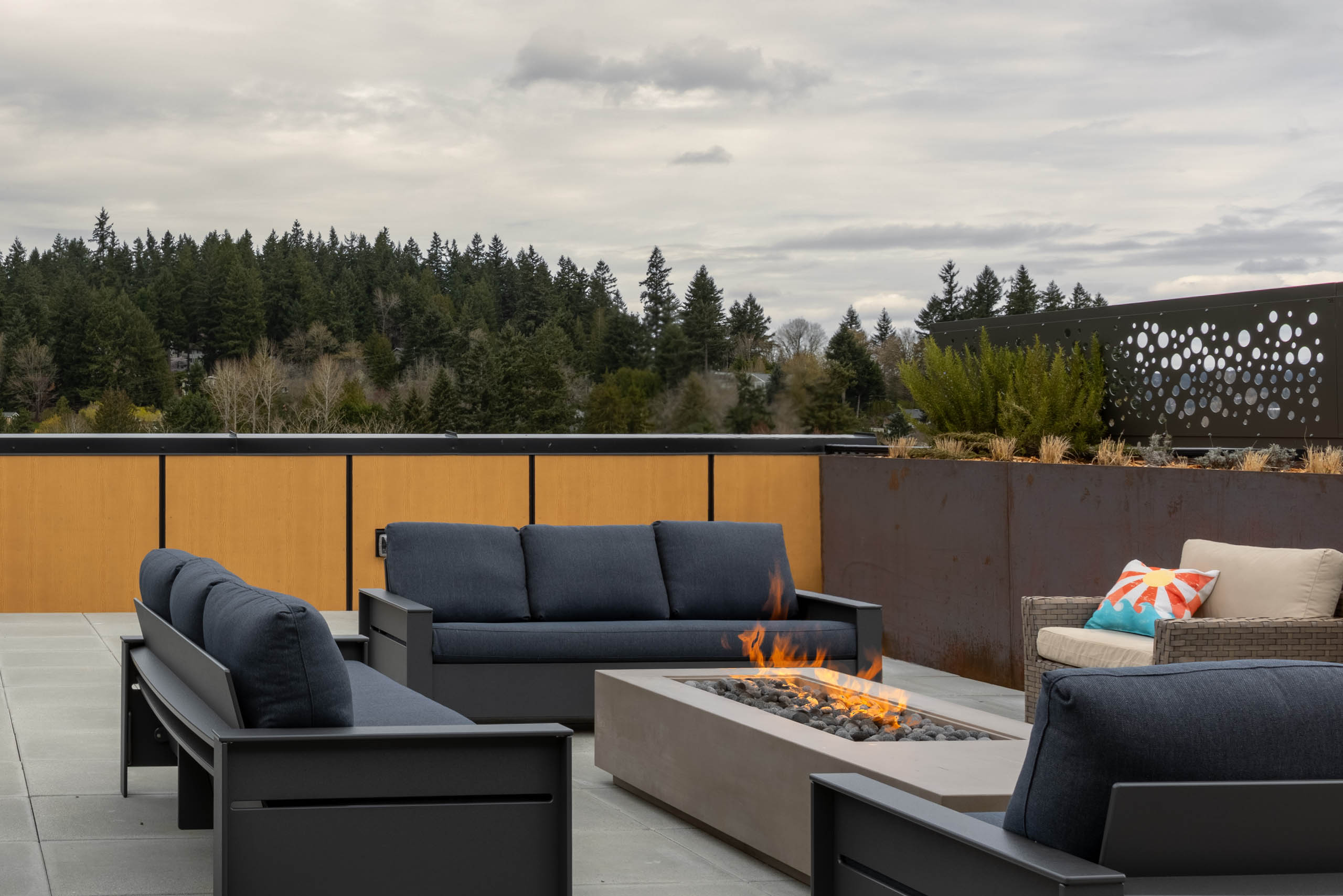 Gray three-piece chairs and a single chair surround an open firepit on a rooftop terrace.