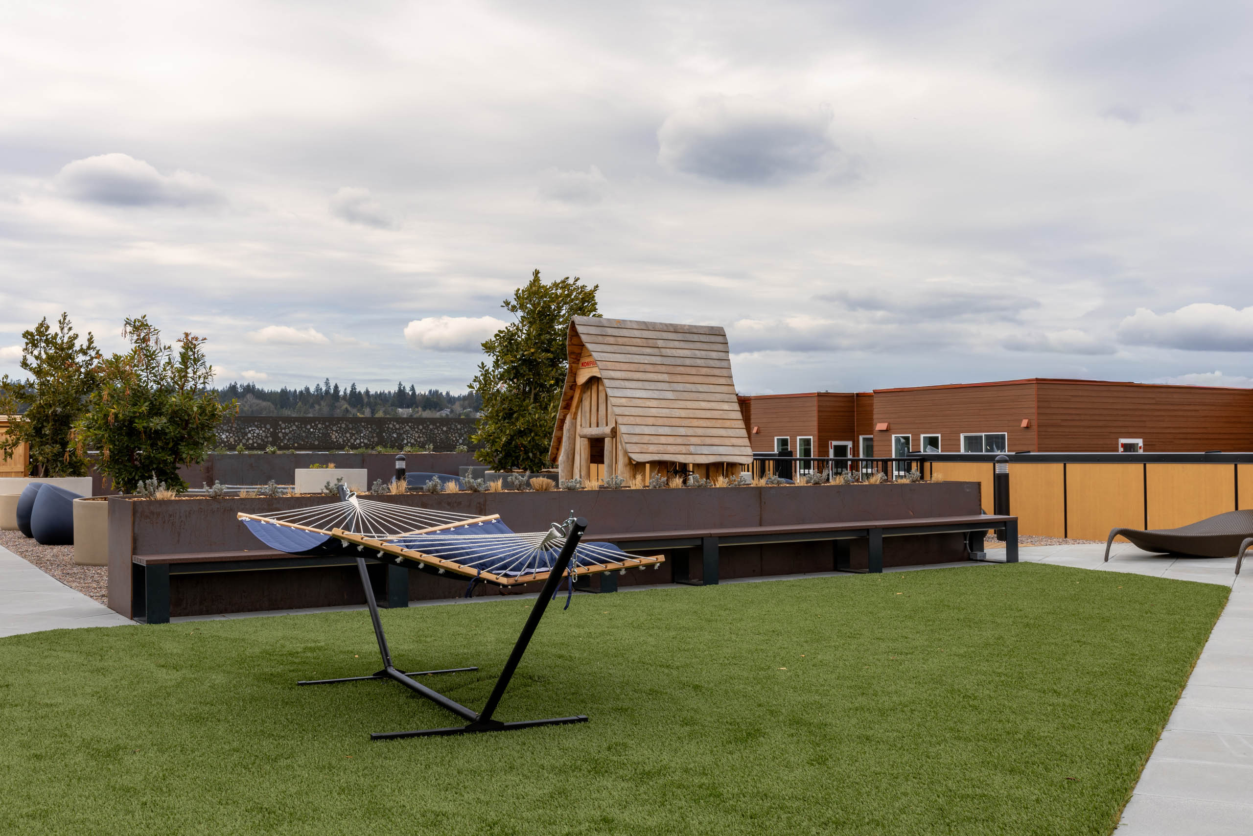 Blue hammock on a rooftop terrace turf patch with seating behind and a small wooden playhouse.