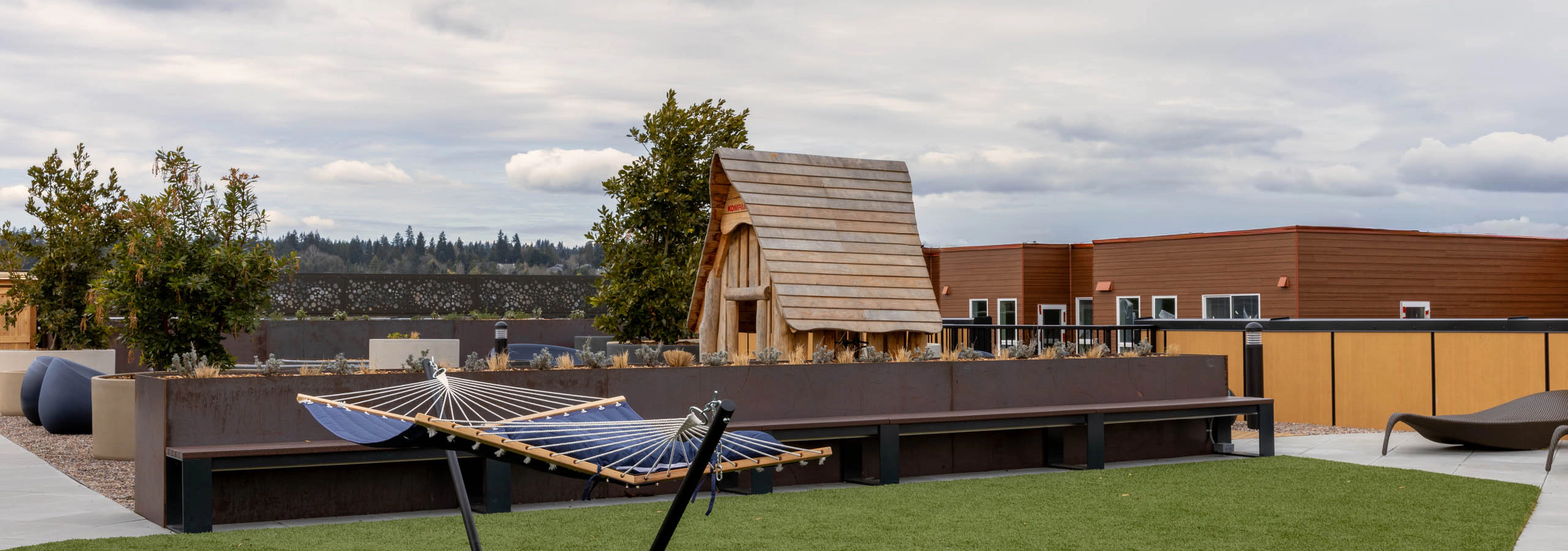 Blue hammock on a rooftop terrace turf patch with seating behind and a small wooden playhouse.
