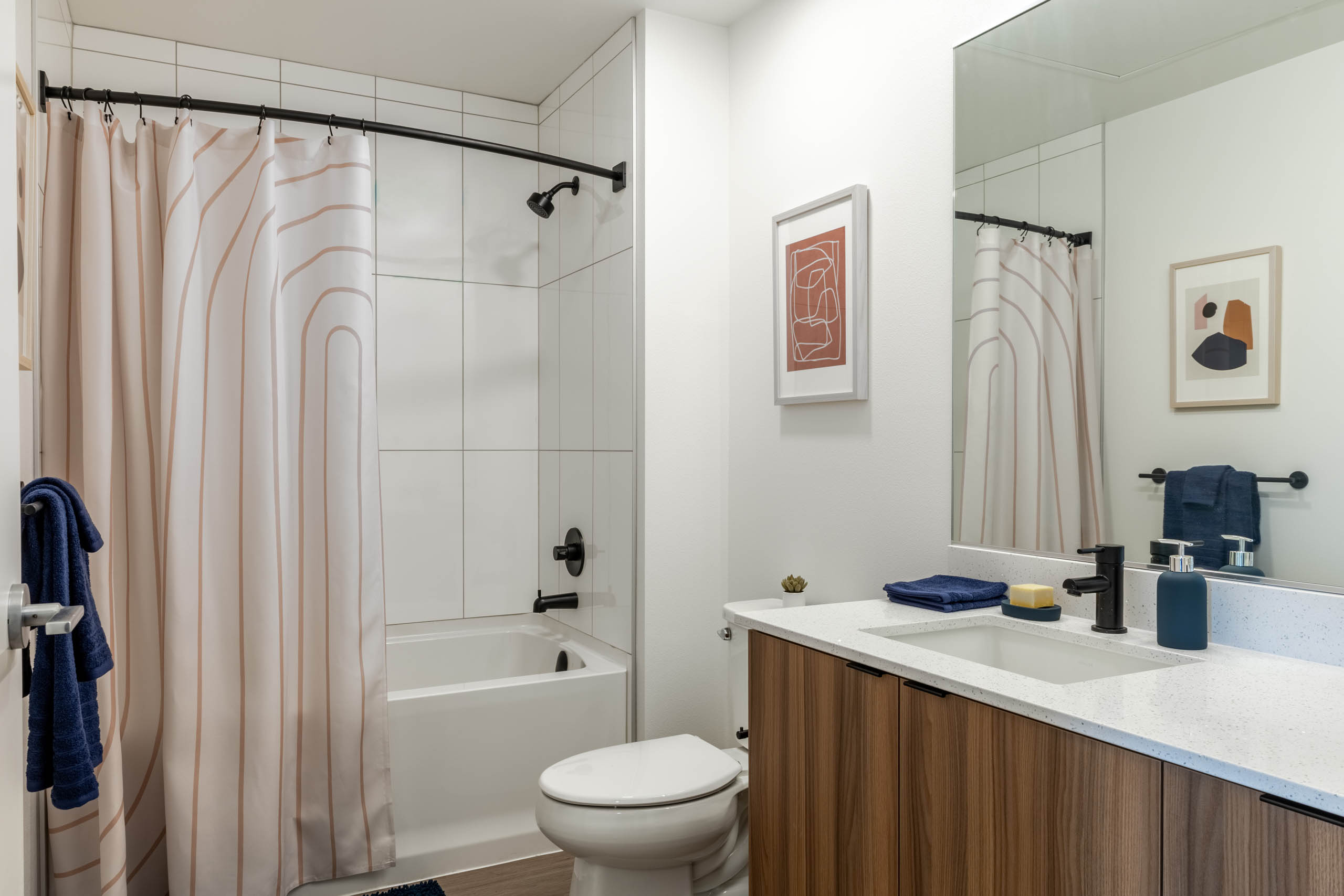 Bright bathroom with white walls, white toilet, next to wooden cabinetry with white sink countertop. White bathtub covered by a cream shower curtain.