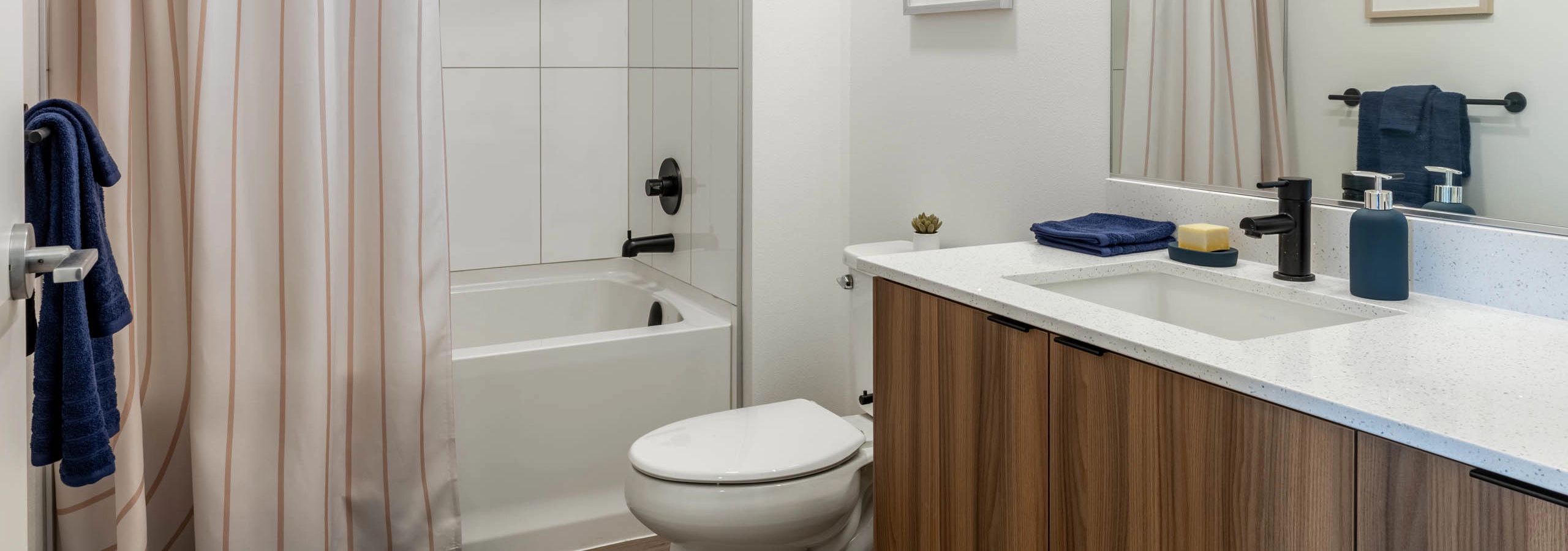 Bright bathroom with white walls, white toilet, next to wooden cabinetry with white sink countertop. White bathtub covered by a cream shower curtain.