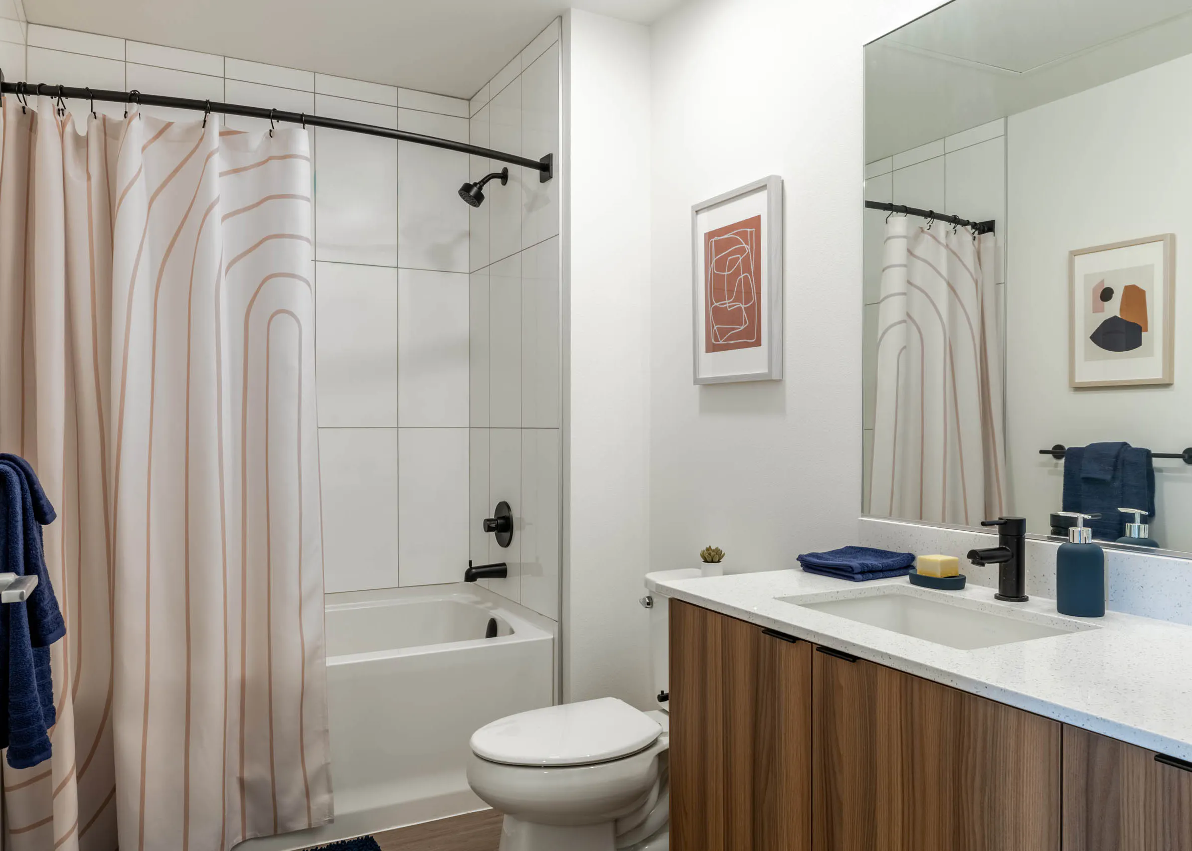 Bright bathroom with white walls, white toilet, next to wooden cabinetry with white sink countertop. White bathtub covered by a cream shower curtain.