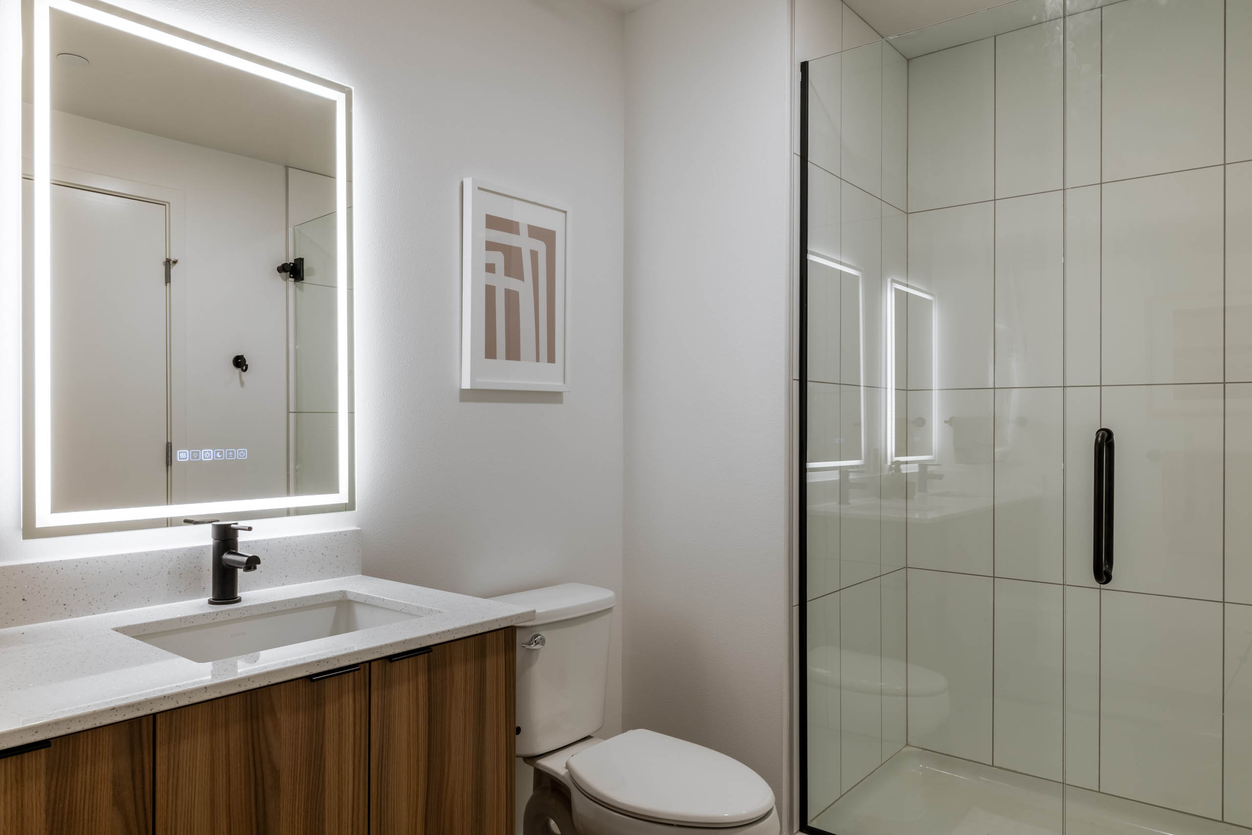 Bright bathroom with white walls, white toilet, next to wooden cabinetry with white sink countertop, light-up mirror and glass shower doors.