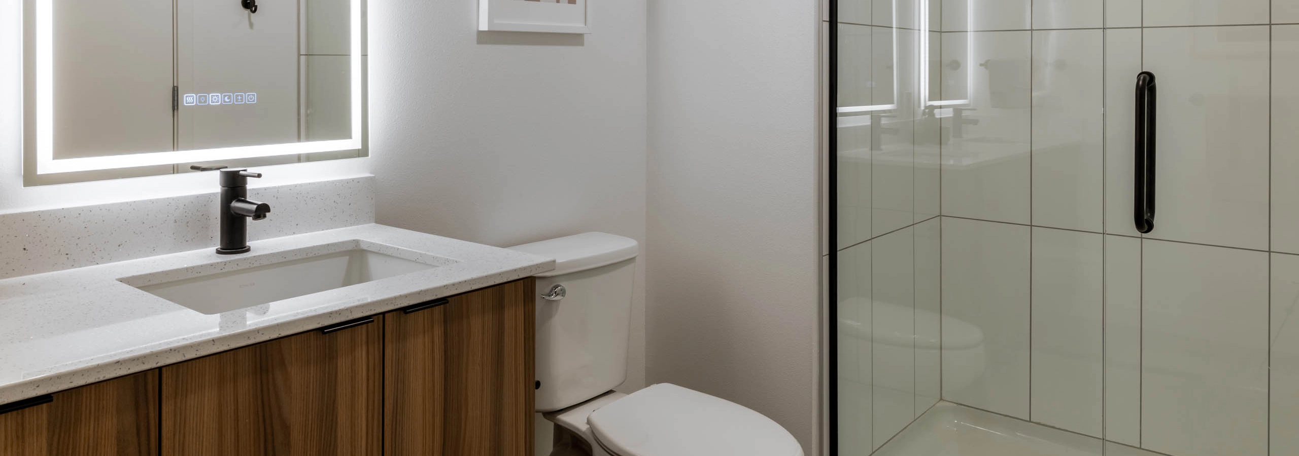 Bright bathroom with white walls, white toilet, next to wooden cabinetry with white sink countertop, light-up mirror and glass shower doors.