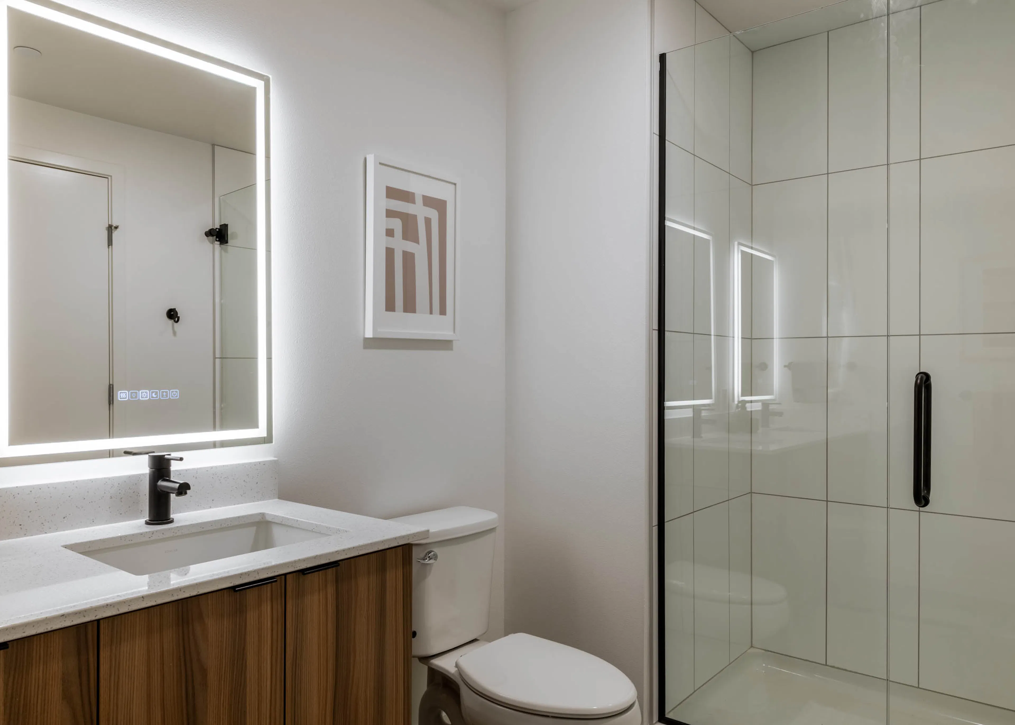 Bright bathroom with white walls, white toilet, next to wooden cabinetry with white sink countertop, light-up mirror and glass shower doors.