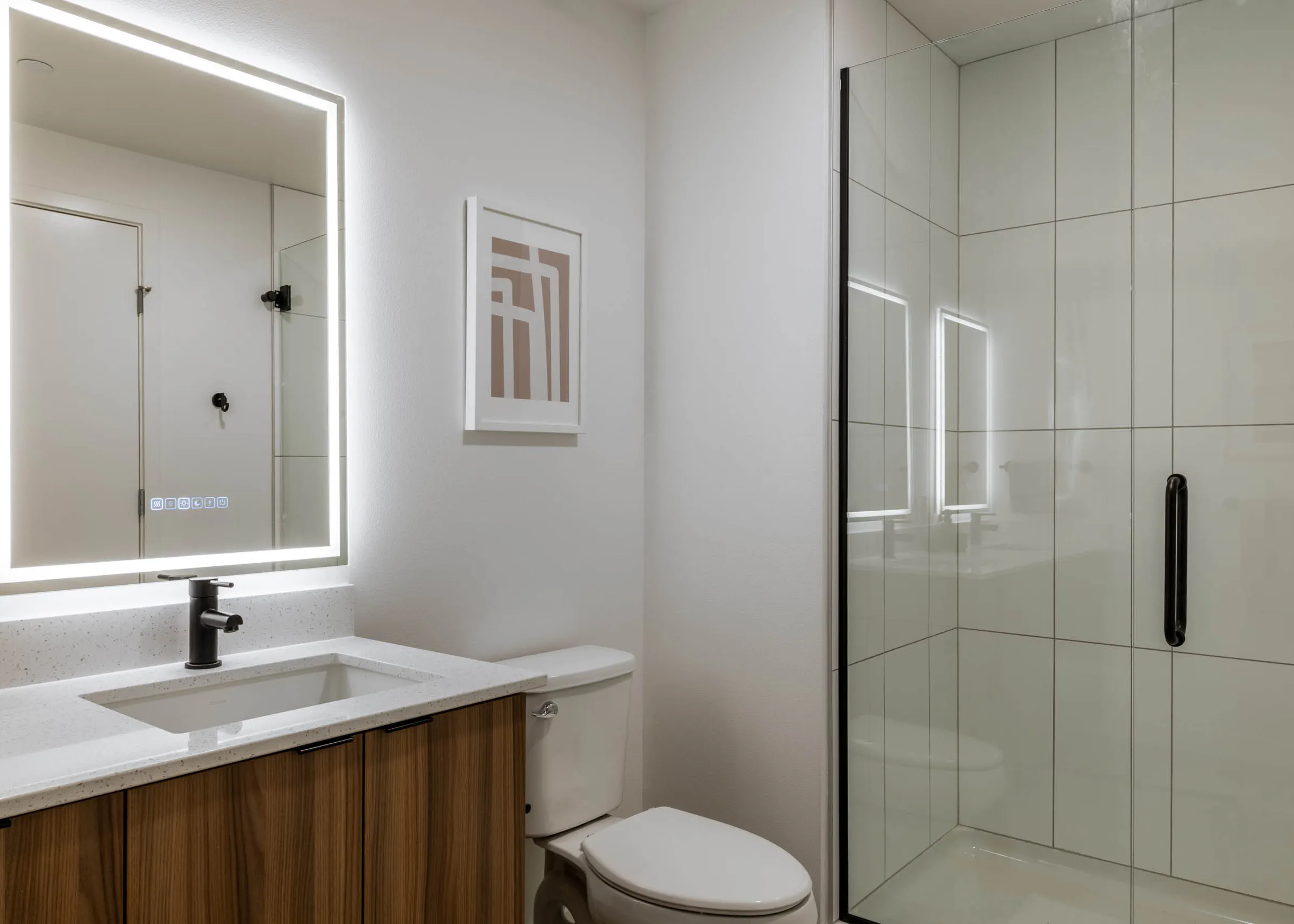 Bright bathroom with white walls, white toilet, next to wooden cabinetry with white sink countertop, light-up mirror and glass shower doors.