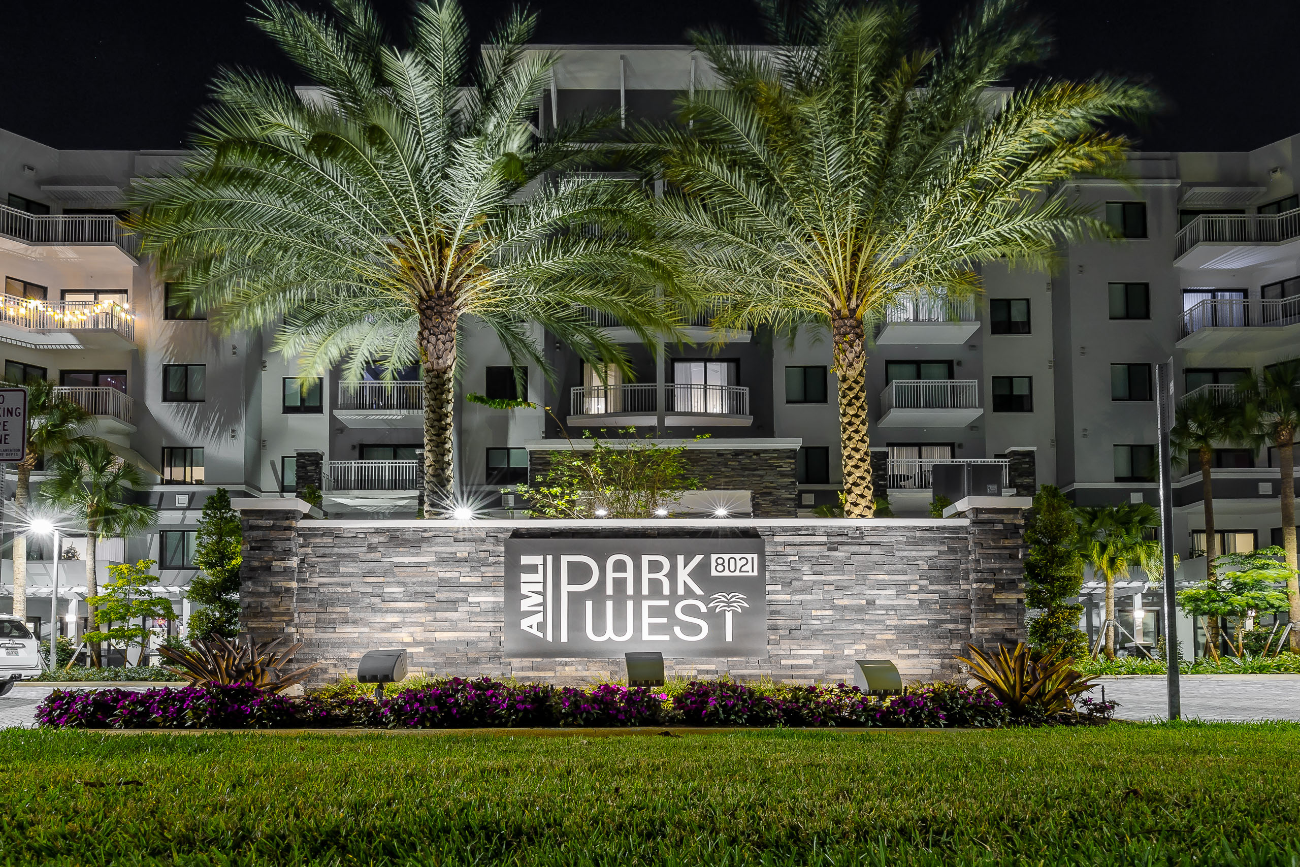 Nighttime view of AMLI Park West apartment building lit monument sign with 2 large palm trees behind it and flowers in front.