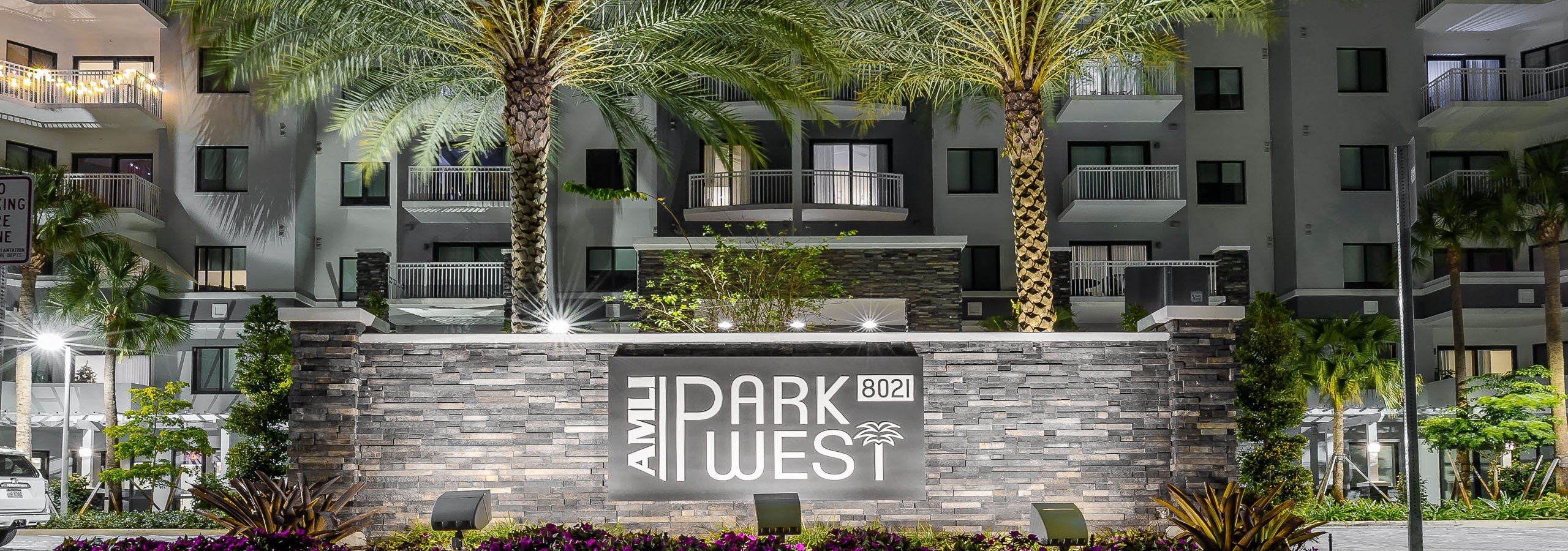 Nighttime view of AMLI Park West apartment building lit monument sign with 2 large palm trees behind it and flowers in front.