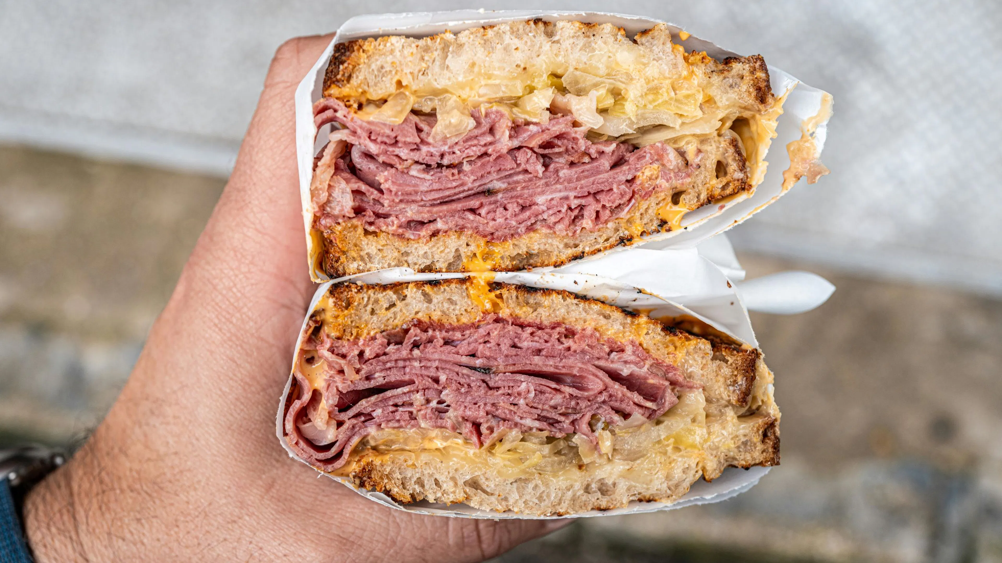 Stacked pastrami sandwich on house-baked bread with melted cheese and sauerkraut, held for a close-up cross-section view.
