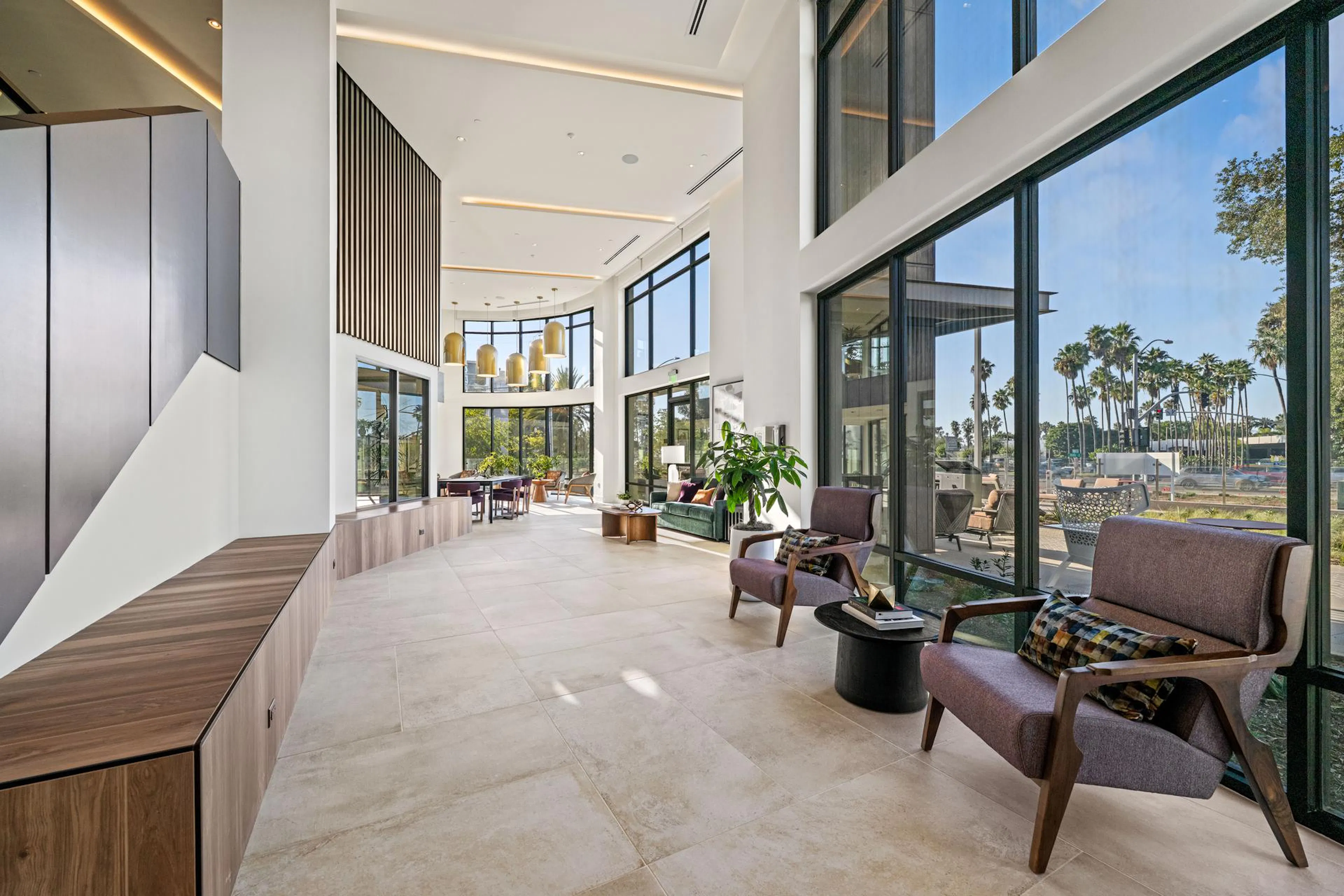 A spacious, high-ceilinged apartment building lobby featuring expansive, double-height floor-to-ceiling windows that look out onto palm trees. The contemporary space is styled with light tile flooring, wood-paneled accent walls, and recessed lighting. In the foreground, two mid-century modern armchairs with textured purple upholstery sit next to a small black side table. The background opens into a longer lounge area adorned with cylindrical gold pendant lights and various seating arrangements.