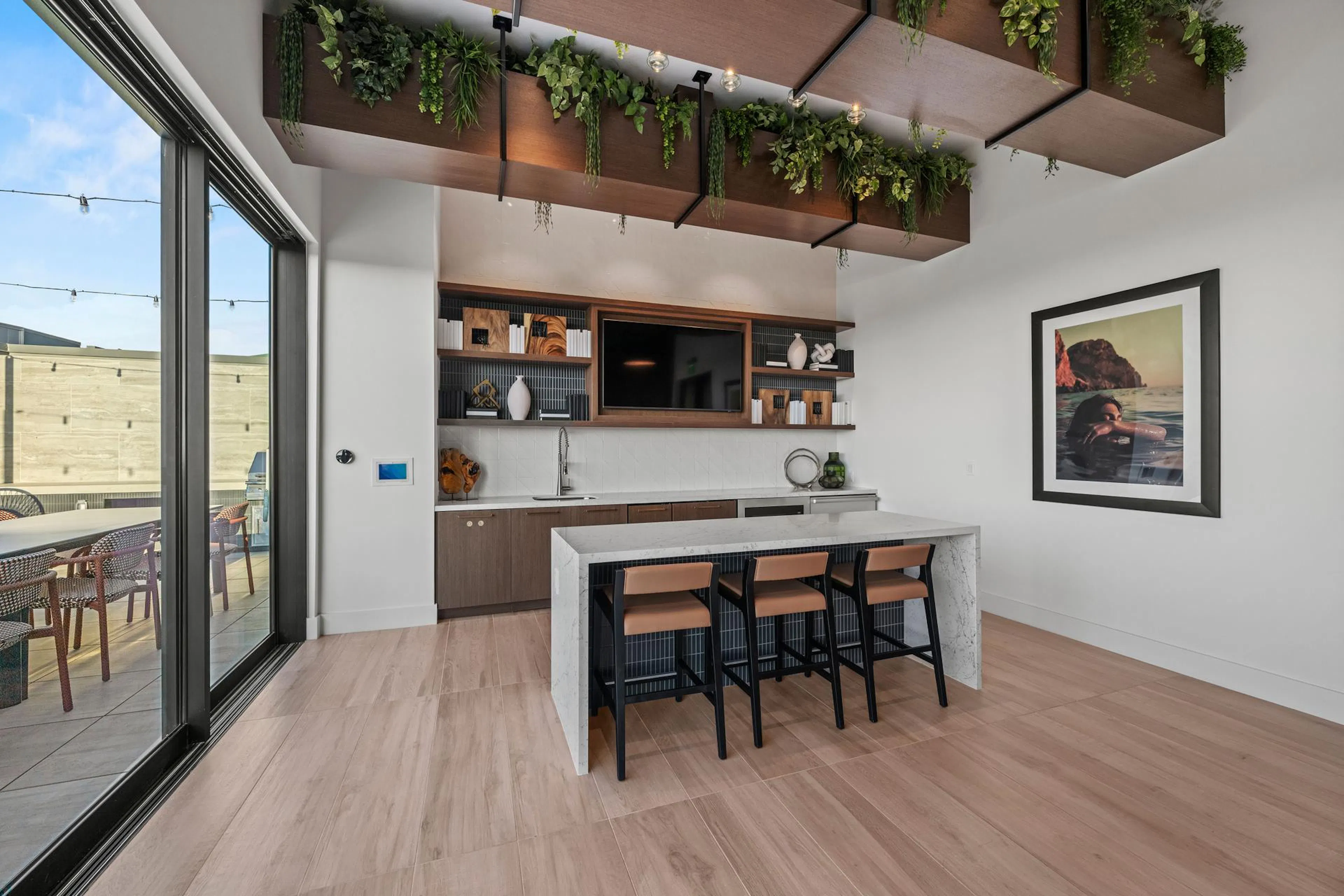A modern resident bar area inside a clubroom featuring a white quartz waterfall island with three black-framed barstools with tan leather cushions. The back bar includes dark wood lower cabinets, a white tile backsplash, a small sink, and open shelving decorated with books and vases framing a wall-mounted flat-screen television. Hanging from the ceiling are large wooden architectural beams overflowing with cascading faux greenery, and a large sliding glass door to the left opens onto a patio