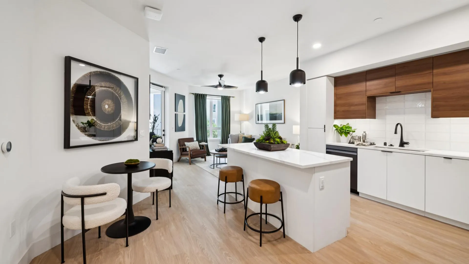 Bright, open-concept apartment interior showing a modern kitchen that flows into a living room. The kitchen features white lower cabinetry, warm wood upper cabinets, a white waterfall-edge island, and camel-colored barstools. In the foreground, a small round black bistro table is flanked by two white bouclé textured chairs under a large piece of abstract circular wall art. The living room in the background features green curtains and a leather armchair.