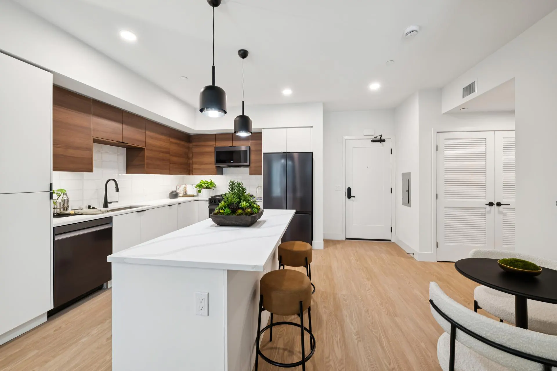 Modern apartment kitchen featuring an island with white quartz countertops and two camel-colored leather barstools. The kitchen has a mix of flat-panel white lower cabinets and warm wood upper cabinets, black appliances, a black faucet, and two matte black pendant lights hanging above the island. In the background, a white front door and a louvered double-door closet are visible against light wood-plank flooring.