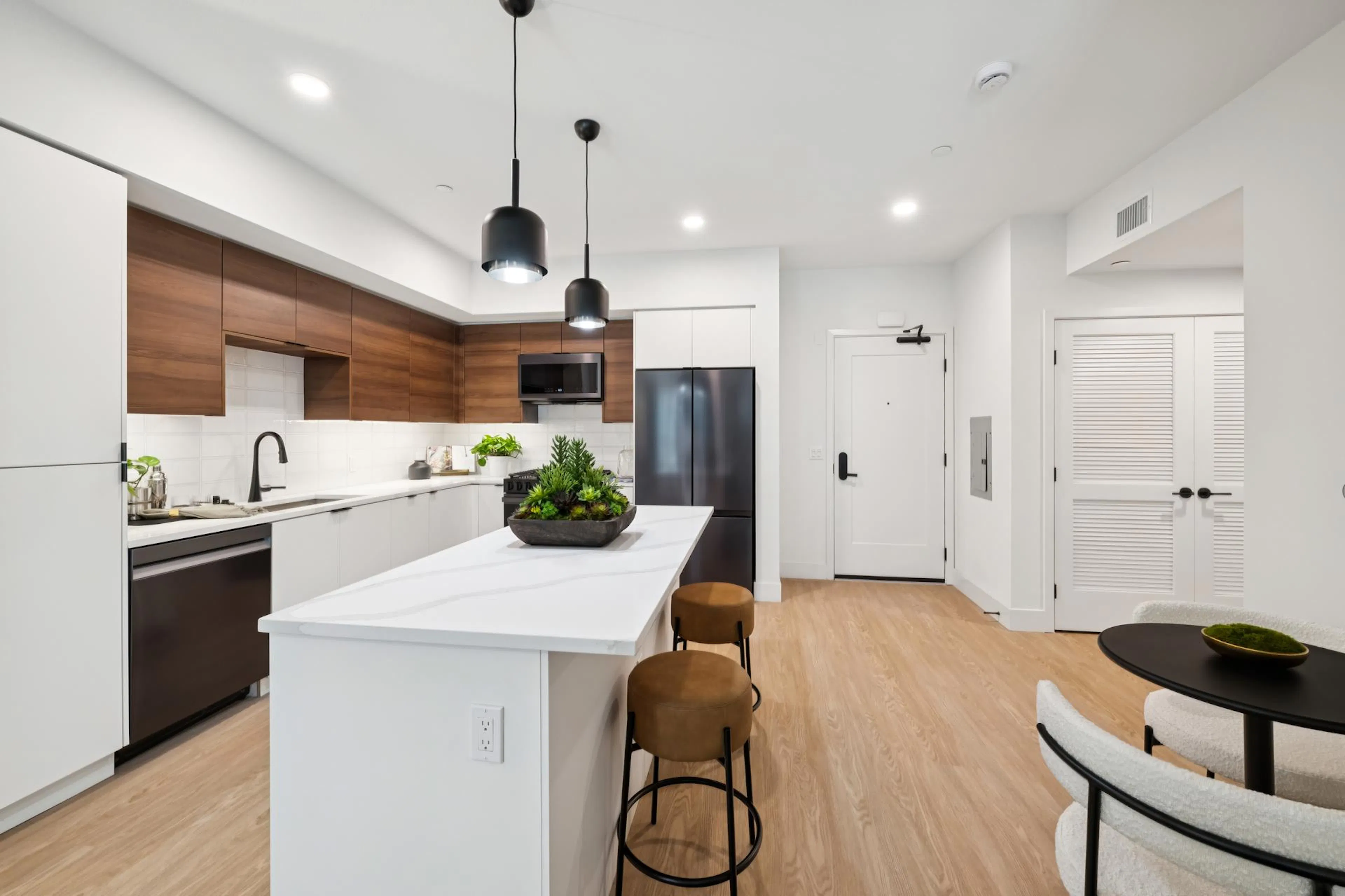 Modern apartment kitchen featuring an island with white quartz countertops and two camel-colored leather barstools. The kitchen has a mix of flat-panel white lower cabinets and warm wood upper cabinets, black appliances, a black faucet, and two matte black pendant lights hanging above the island. In the background, a white front door and a louvered double-door closet are visible against light wood-plank flooring.