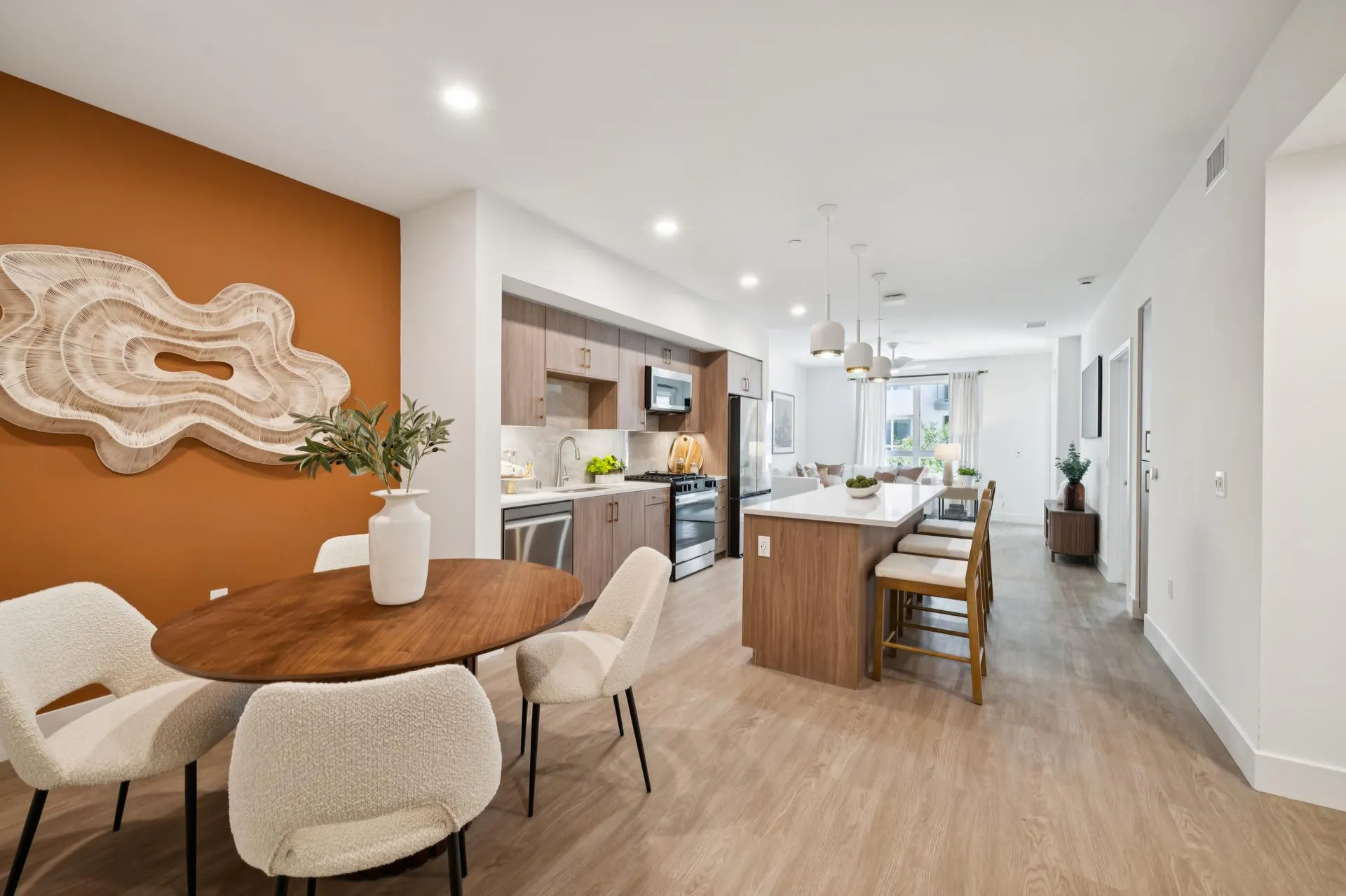 Modern open-plan apartment featuring a dining area with an orange accent wall, a round wooden dining table, and four white textured chairs. A large abstract woven art piece hangs on the accent wall. The adjacent kitchen features light wood cabinets, white countertops, stainless steel appliances, and a central kitchen island with three cushioned barstools leading into a bright living area.