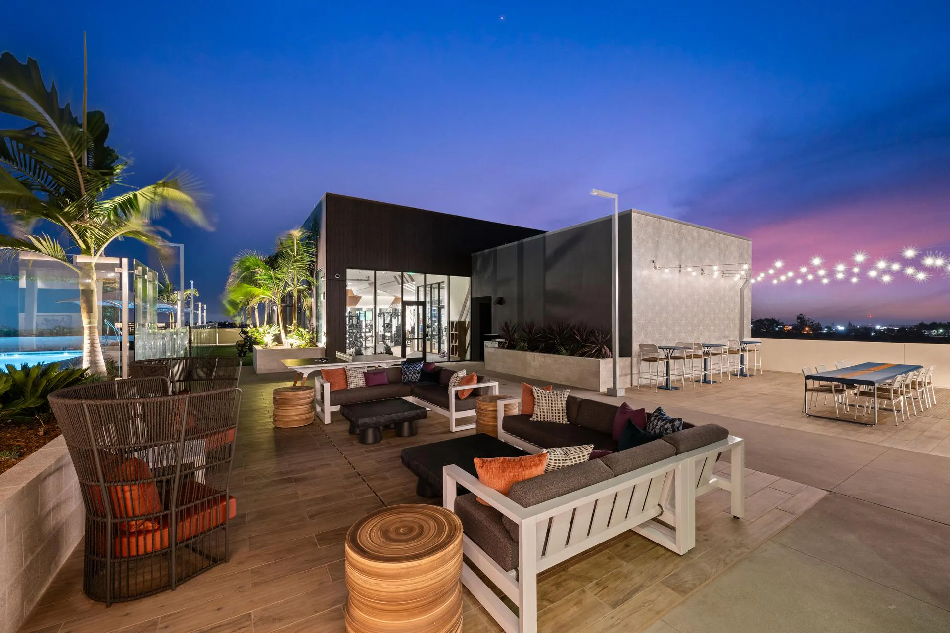Luxury apartment rooftop terrace at dusk with a vibrant purple and blue sunset sky. The expansive patio features multiple outdoor seating arrangements, including gray sectional sofas with orange and purple accent pillows, woven armchairs, and slatted wood-look accent tables. A long dining table with seating sits under warm string lights, and an indoor fitness center with floor-to-ceiling windows is visible in the background next to potted palm trees.