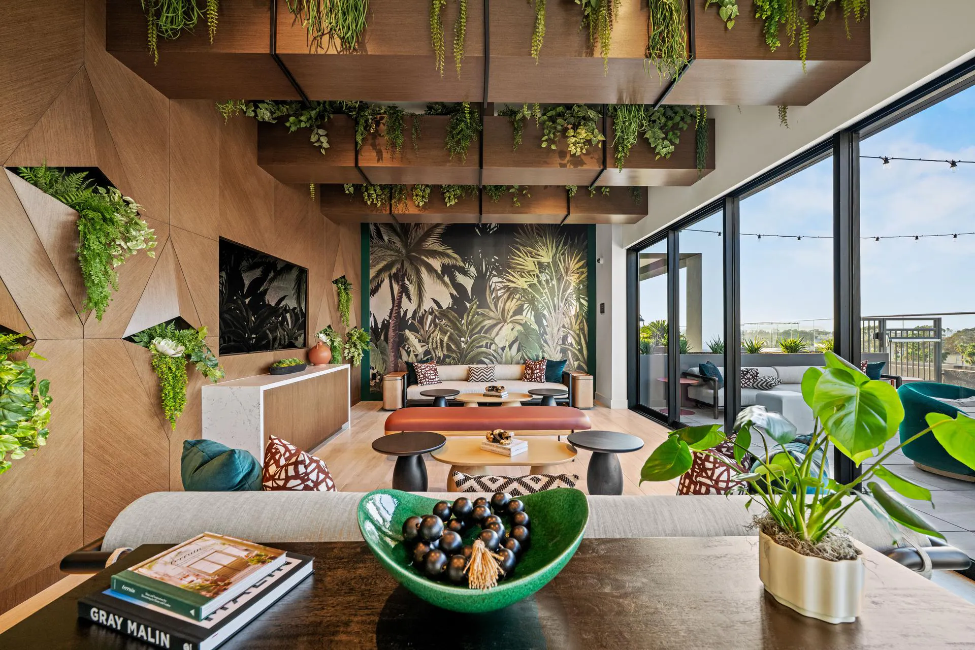 A vibrant apartment resident clubroom featuring a biophilic interior design. The room is decorated with extensive faux greenery spilling from floating wooden ceiling beams and geometric wall cutouts. A large tropical palm tree mural covers the back wall behind a cream-colored sofa. In the foreground, a dark wood table displays a textured green bowl with black beads, looking out toward a large sliding glass door that opens to a rooftop patio.