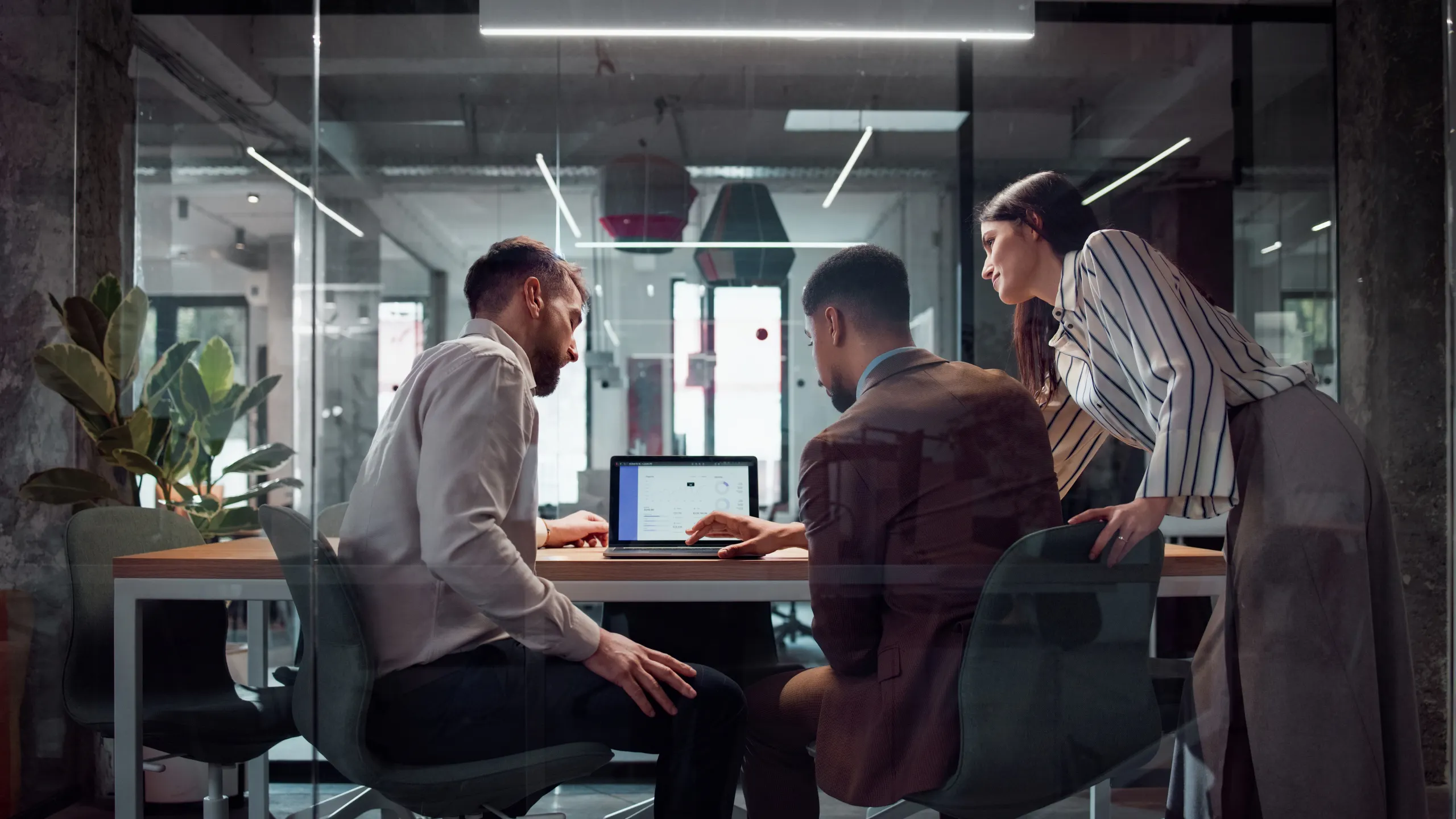 Three work colleagues look at a laptop in a modern conference room with glass walls.