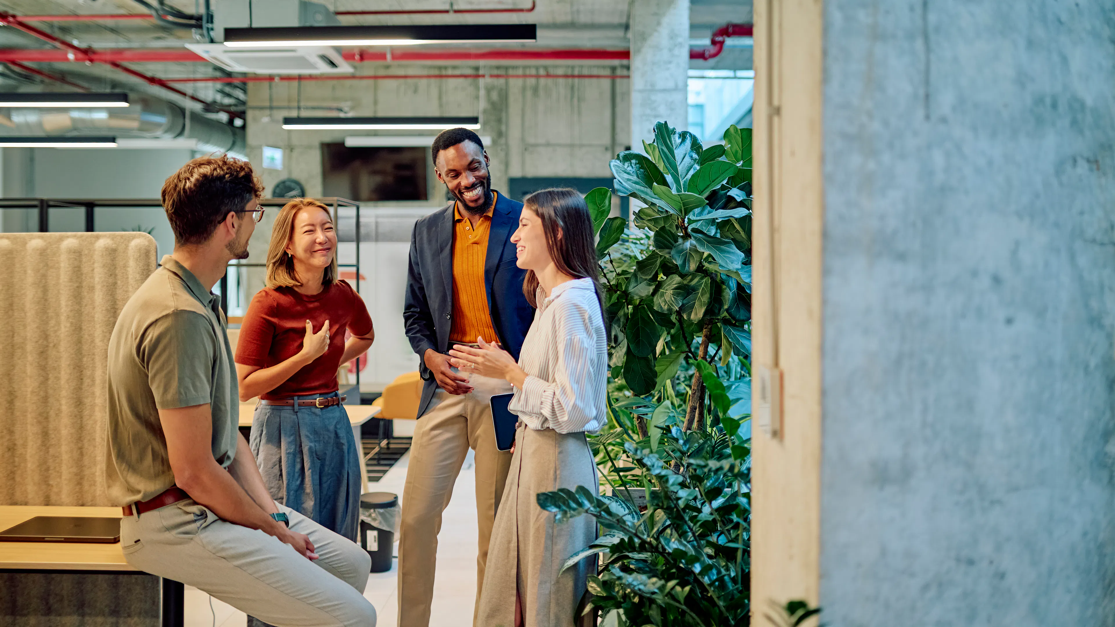 Four coworkers chatting and smiling in a modern open-plan office with exposed concrete ceilings, greenery, and collaborative workspace furnishings.