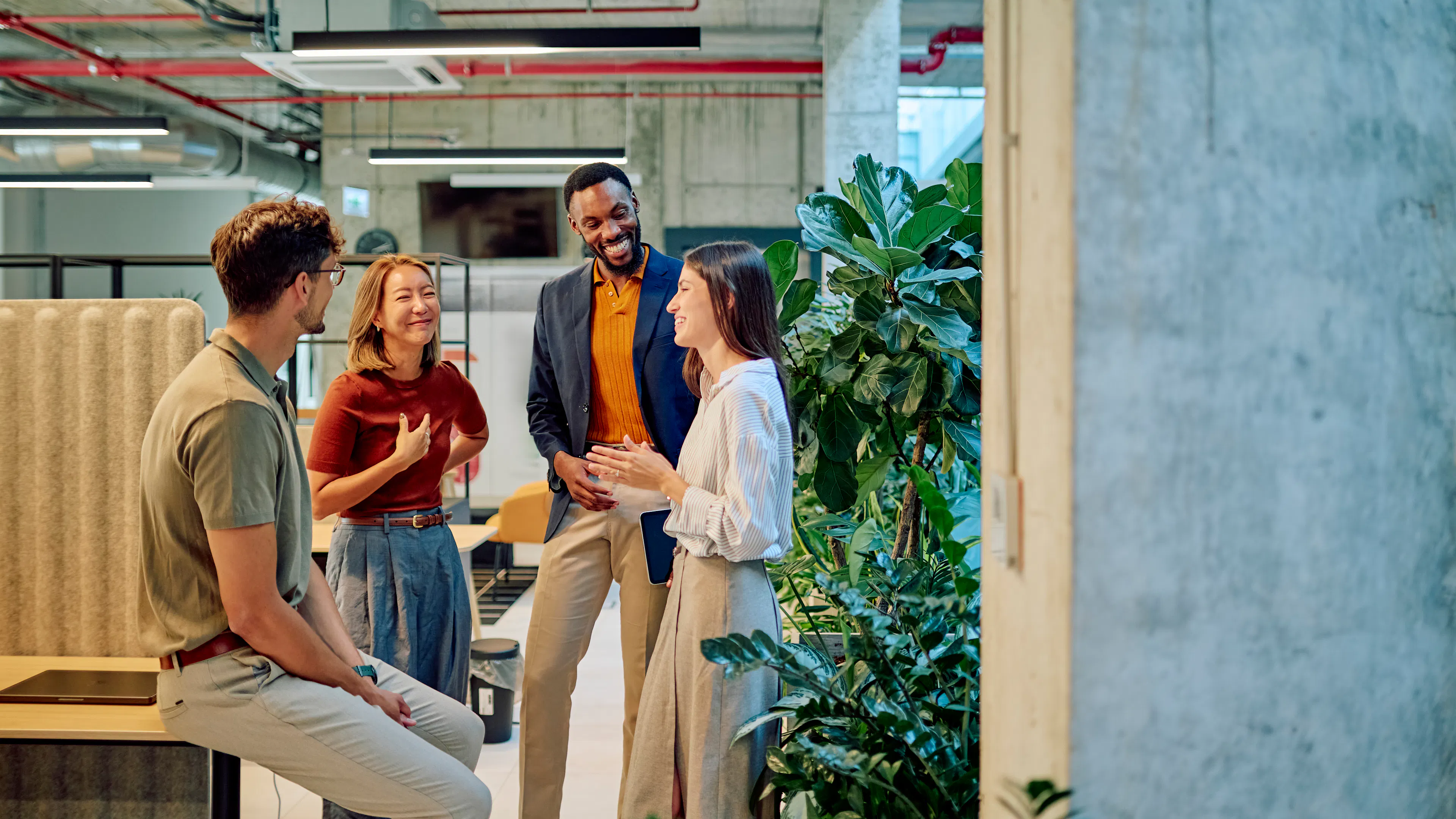 Four coworkers chatting and smiling in a modern open-plan office with exposed concrete ceilings, greenery, and collaborative workspace furnishings.
