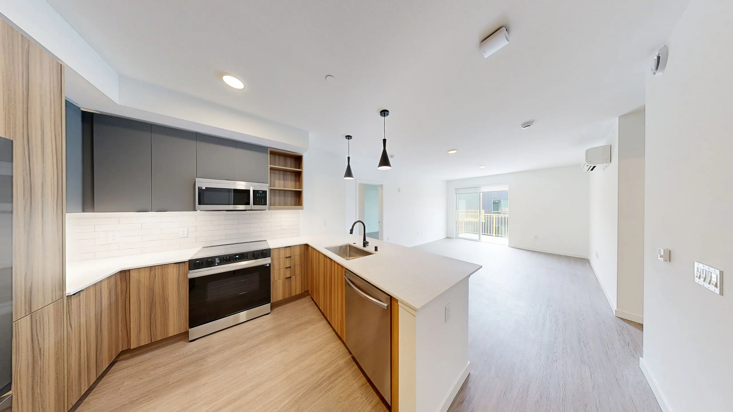A large, naturally lit kitchen and living room apartment space with white walls, light wood style floors, sleek wooden kitchen cabinets and white countertops
