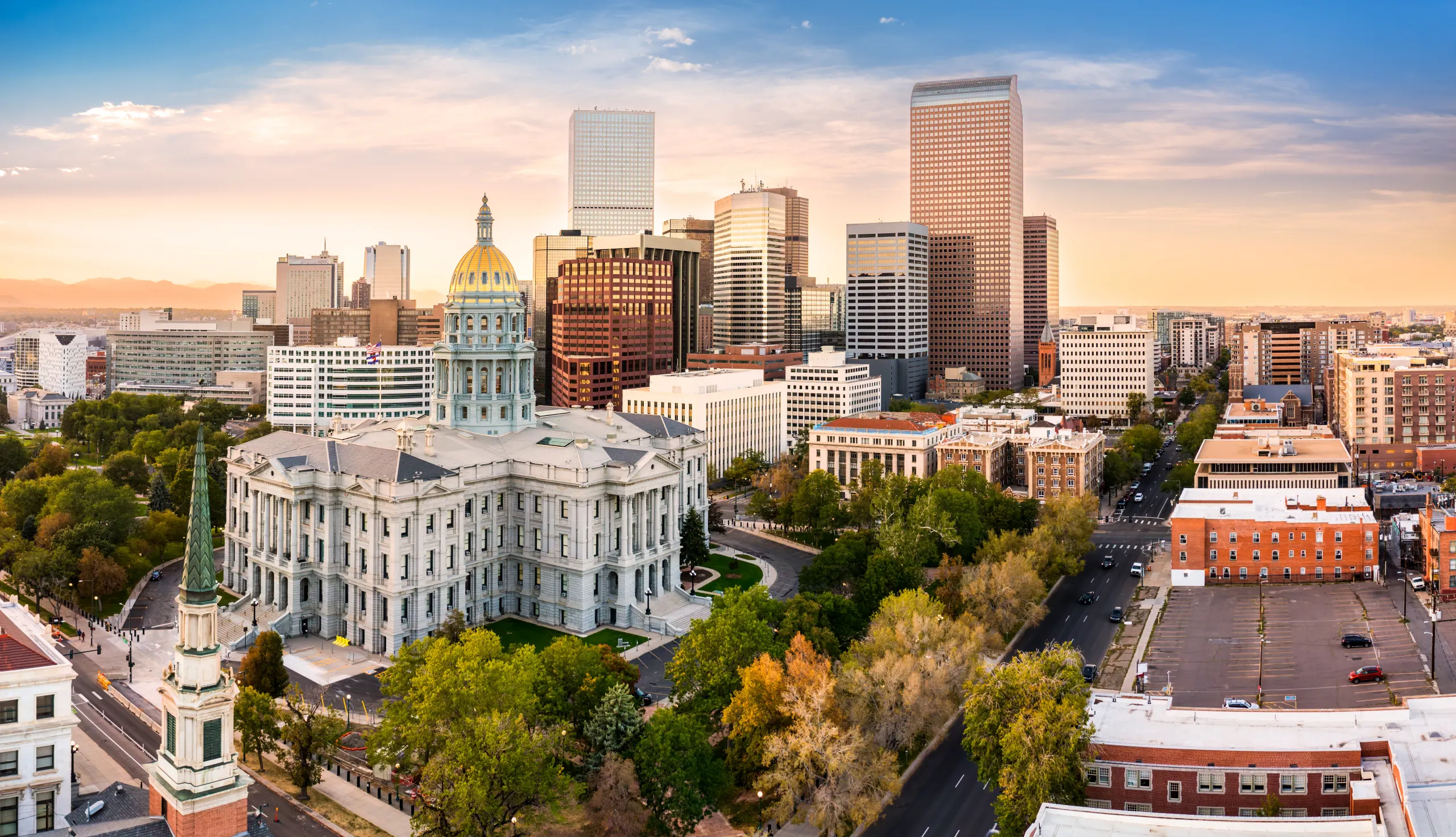 Denver skyline at sunset featuring the Colorado State Capitol building with its golden dome in the foreground surrounded by trees