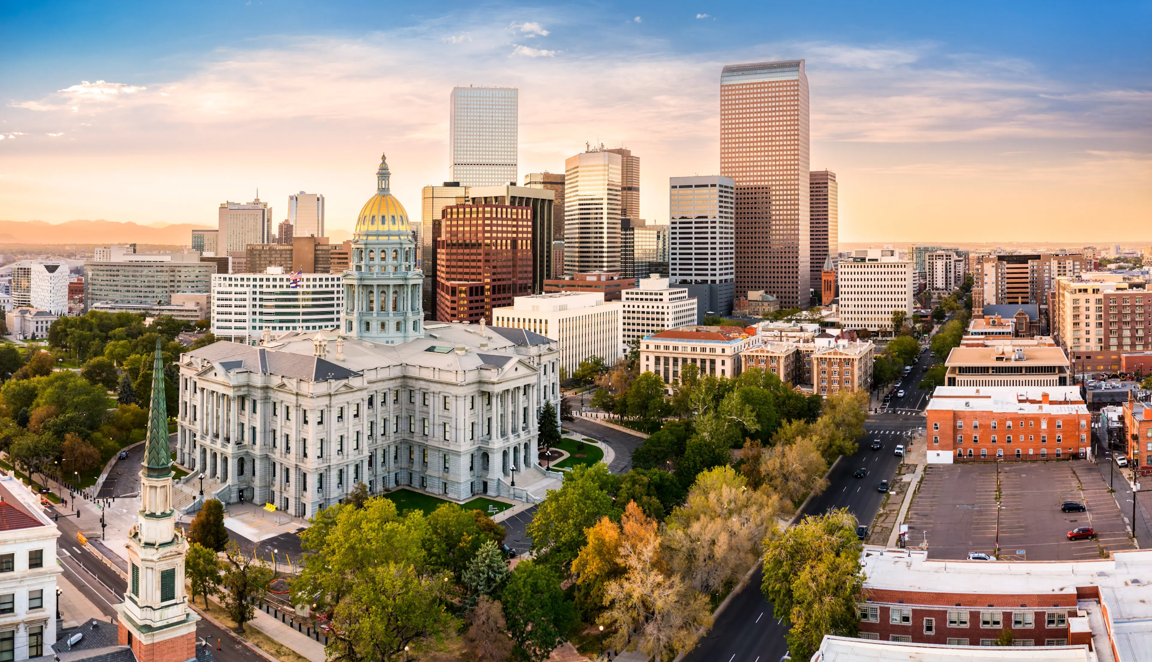 Denver skyline at sunset featuring the Colorado State Capitol building with its golden dome in the foreground surrounded by trees