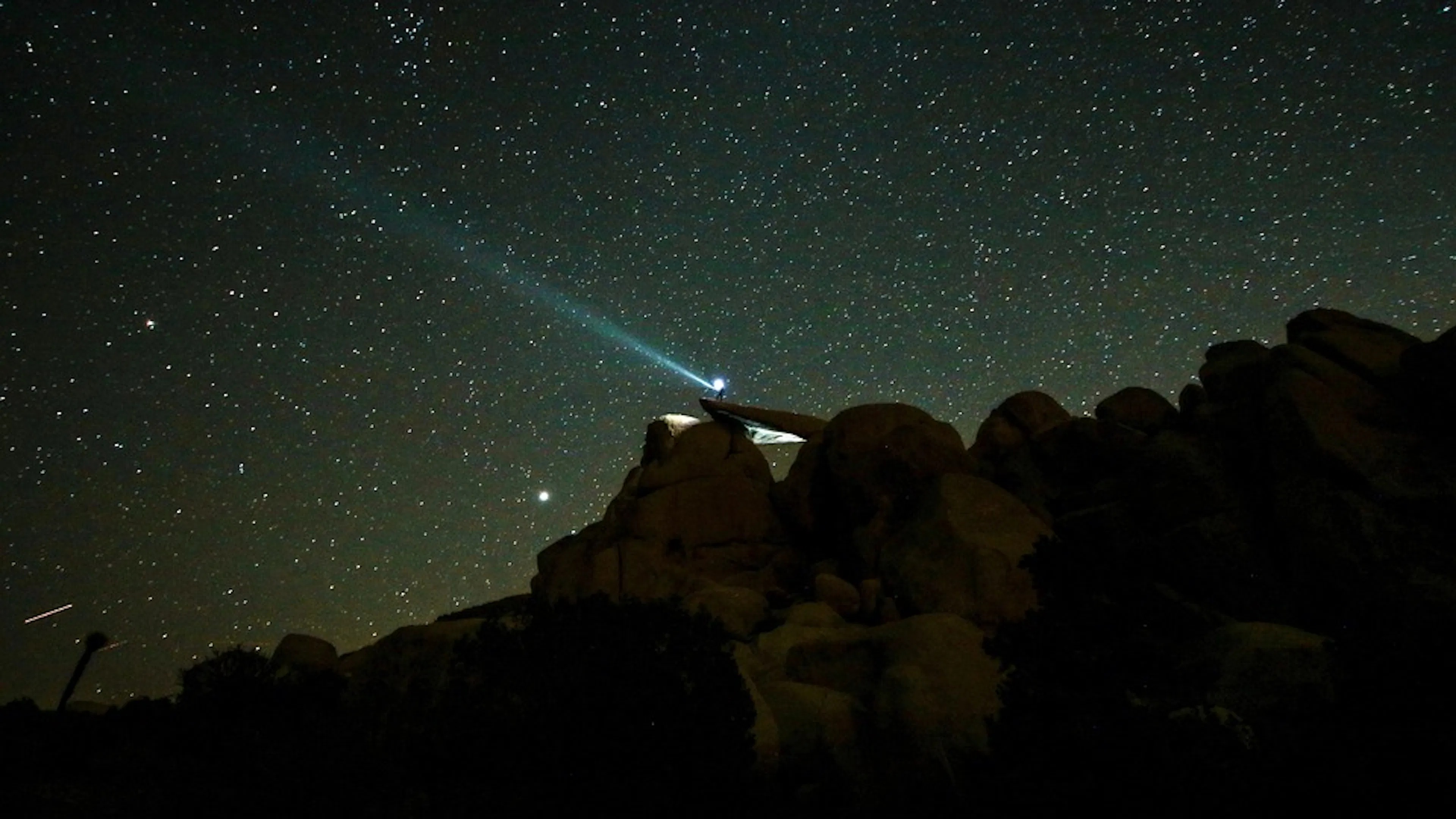 A long exposure shot of a hiker on top of a rocky peak with a flashlight pointing up into a starry sky