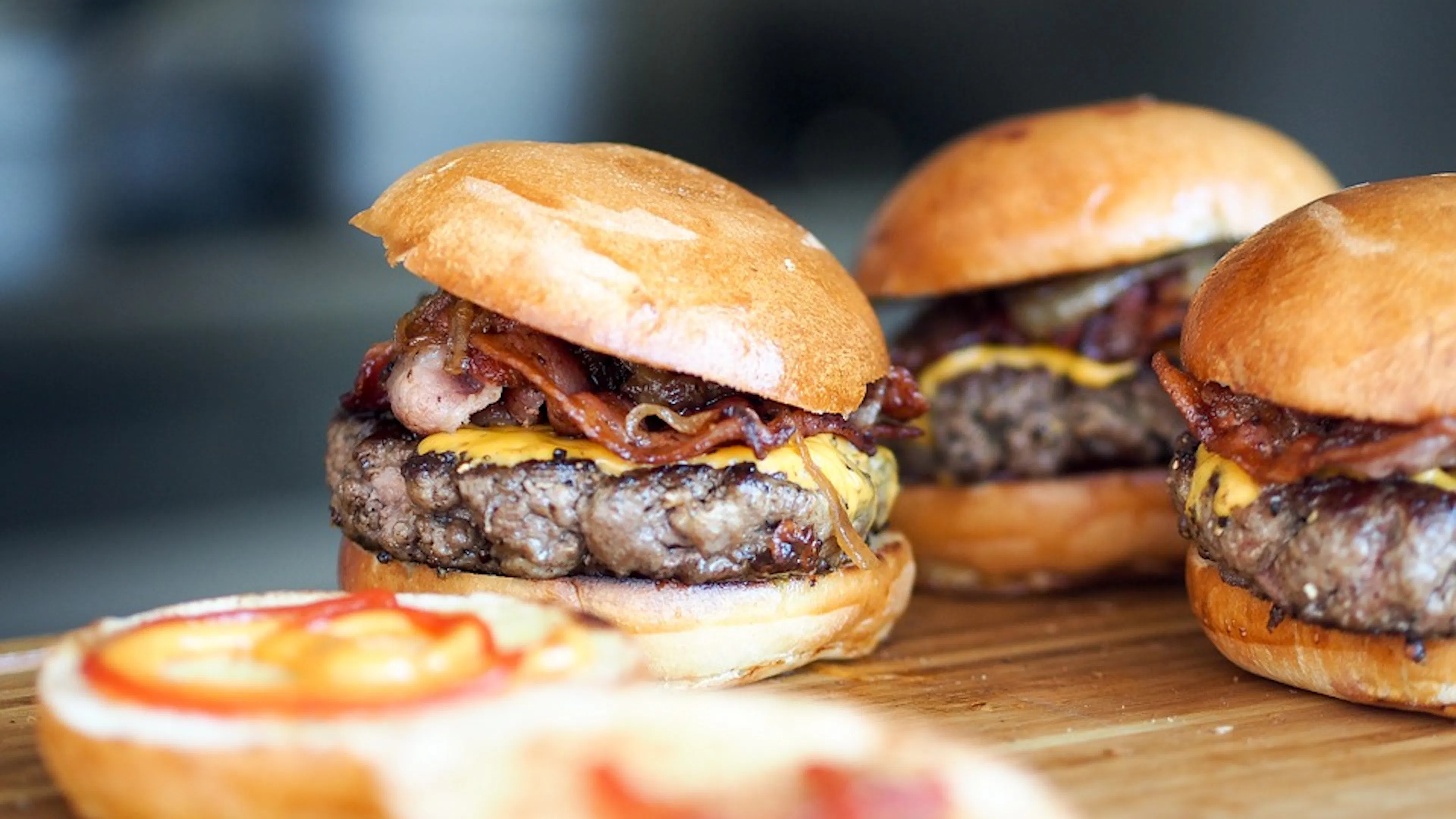 Three freshly made cheeseburgers with crispy bacon on a wooden cutting board and slathered with ketchup