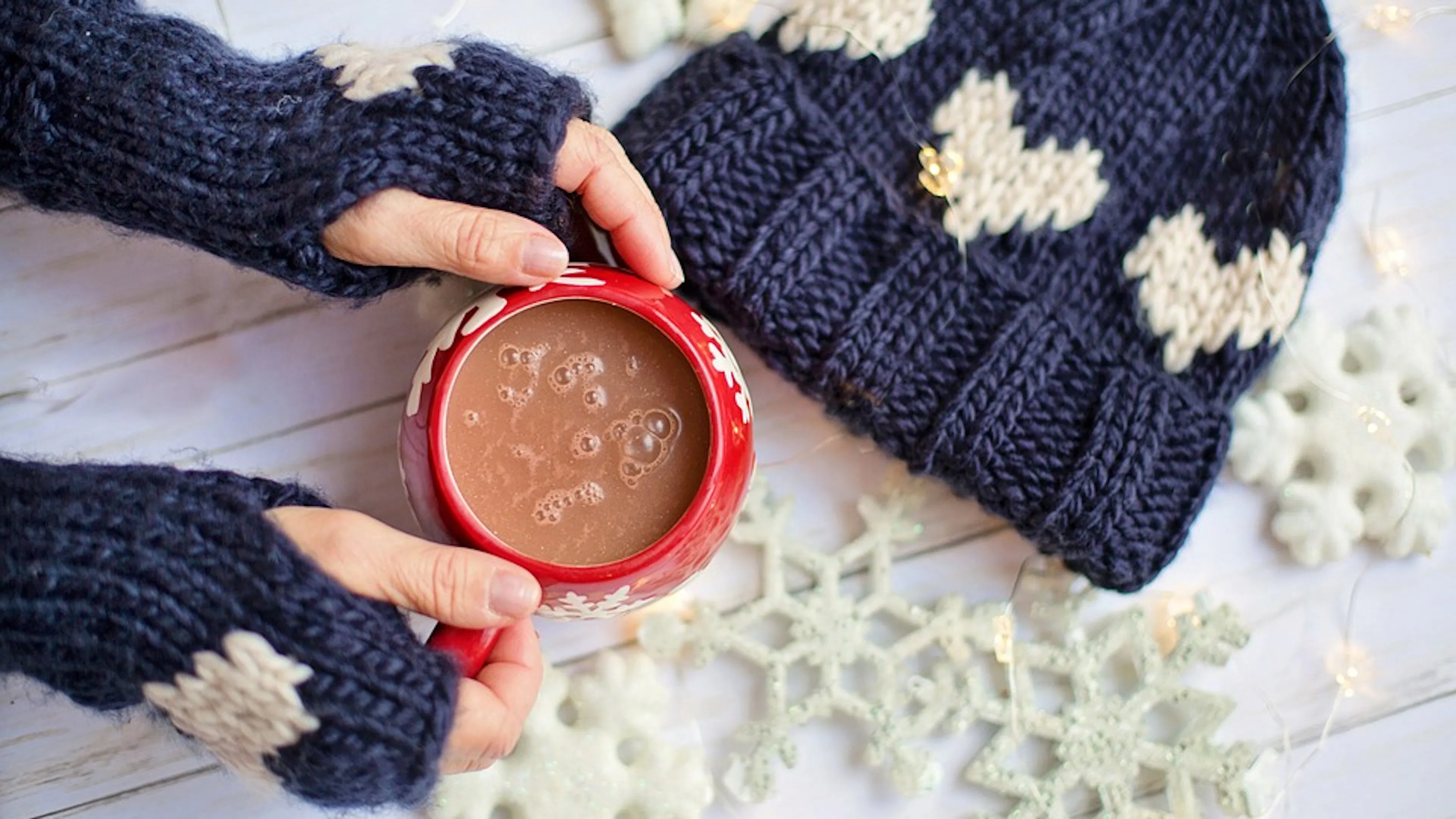 A womans hands holding a red mug of hot cocoa photographed from above with decorative snowflakes scattered around