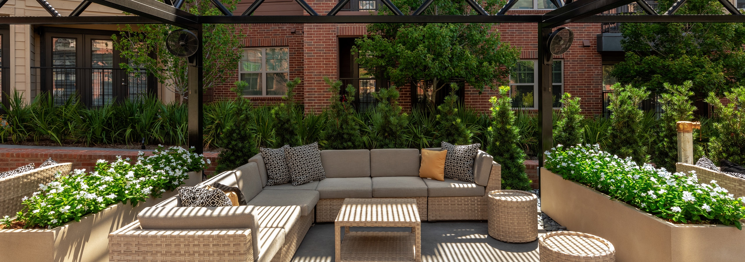Daytime view of resident cabana area with a large wicker L shaped couch and table surrounded by lush greenery and flowers