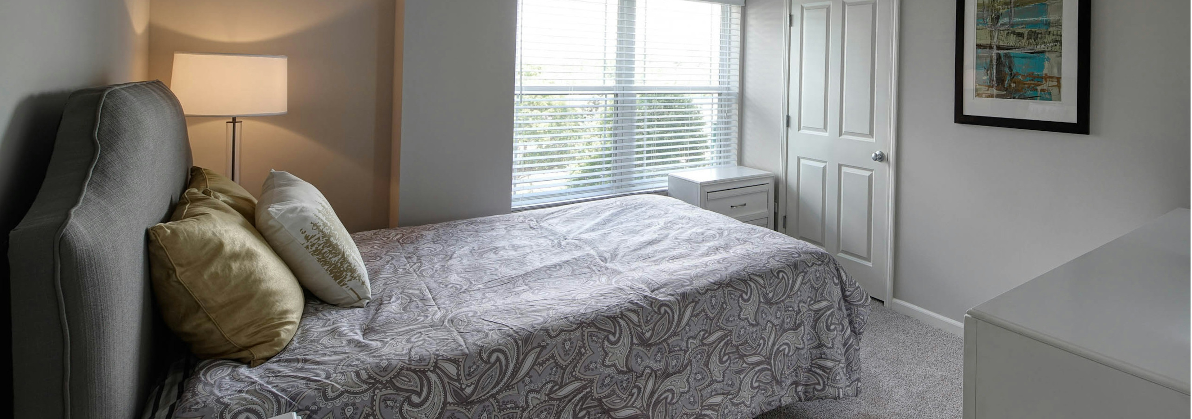 Bedroom at AMLI at Seven Bridges with carpeted floors and grey bedding with a large window showing a bright daytime view