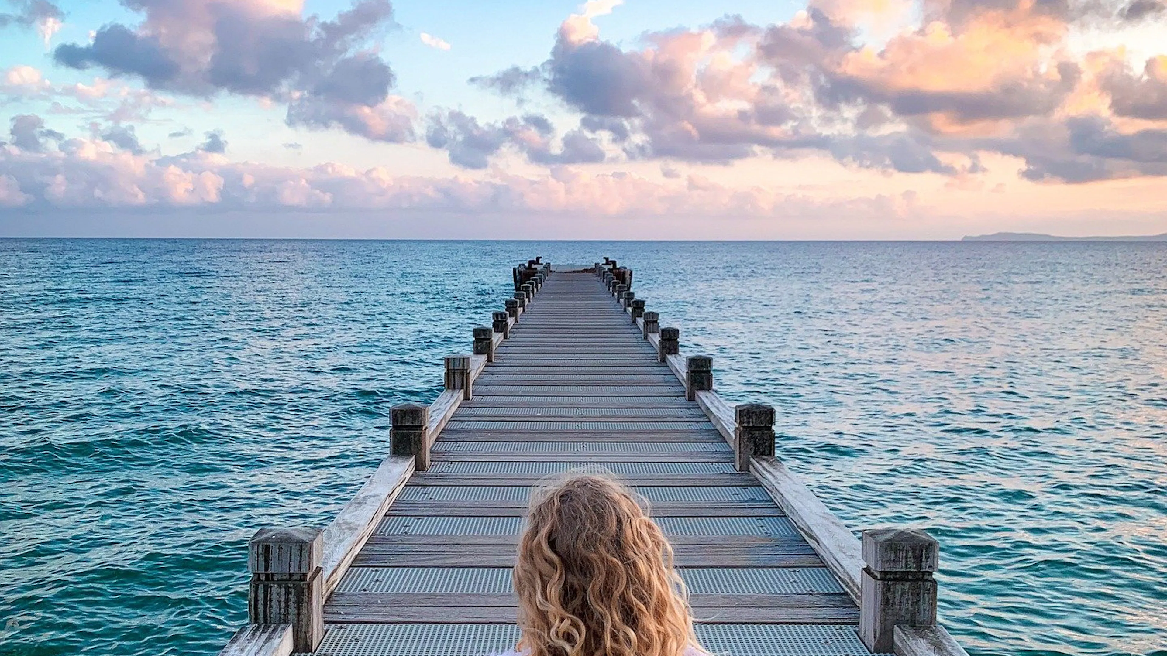 woman with long blonde wavy hair sitting on a boardwalk extending over the ocean