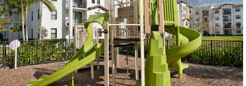 Outdoor green children’s playground at AMLI Doral features stairs for climbing, two slides and all set on rubber floor