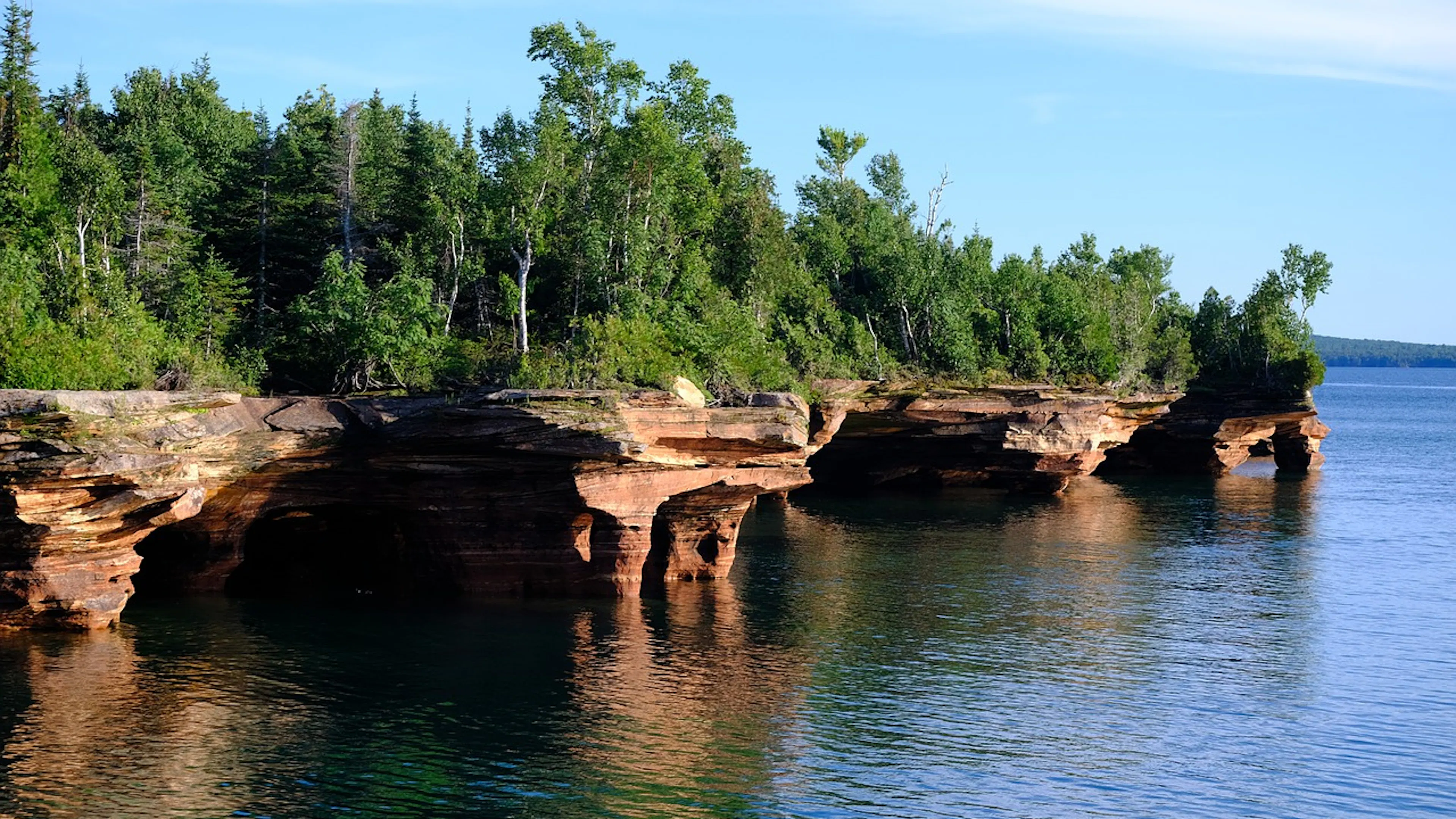 An island with rocky sandstone cliffs and topped with lush forests sticking out of a crystal clear blue lake