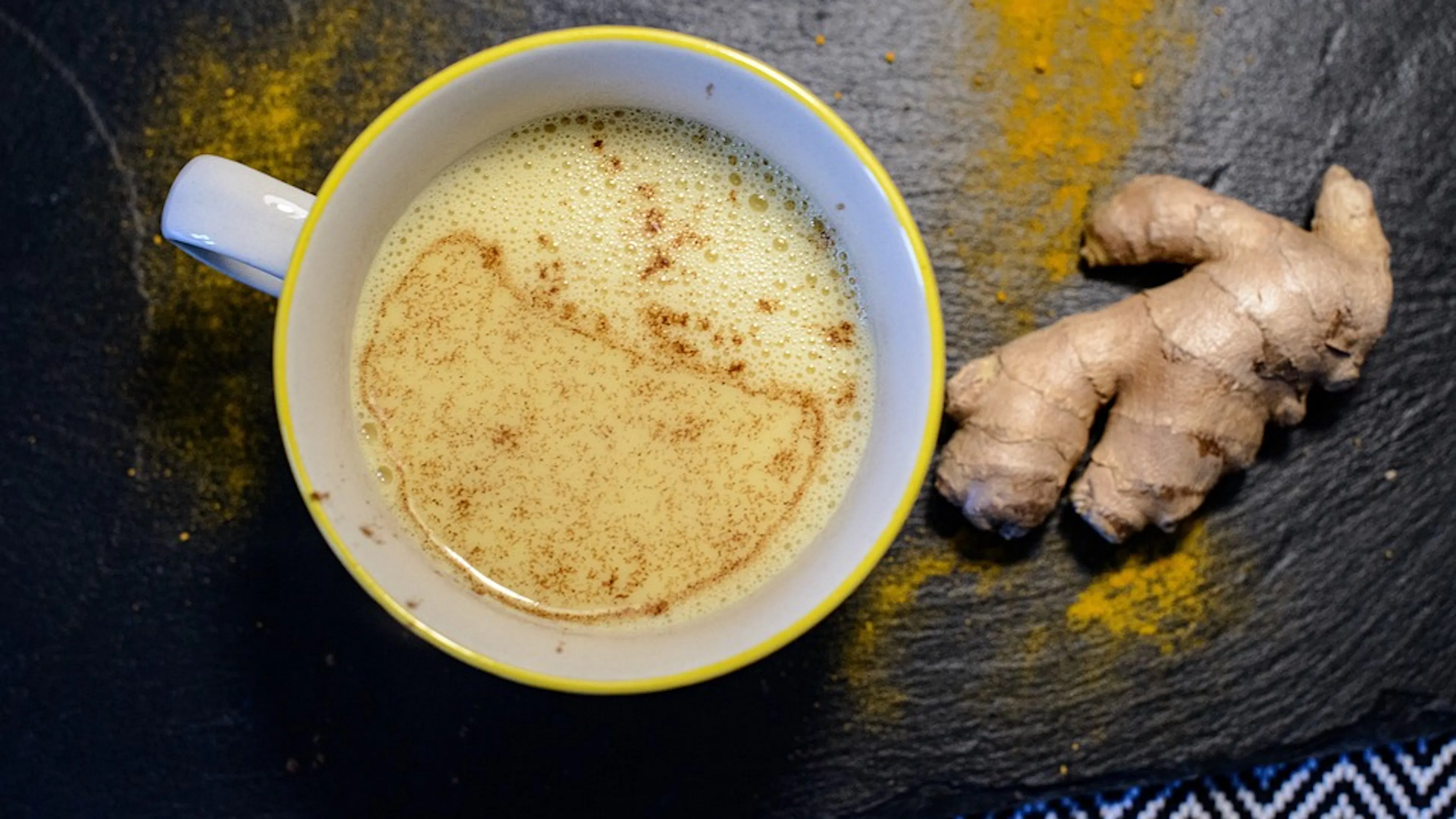 A cup of chai tea next to a piece of ginger root on a dark black stone table photographed from above
